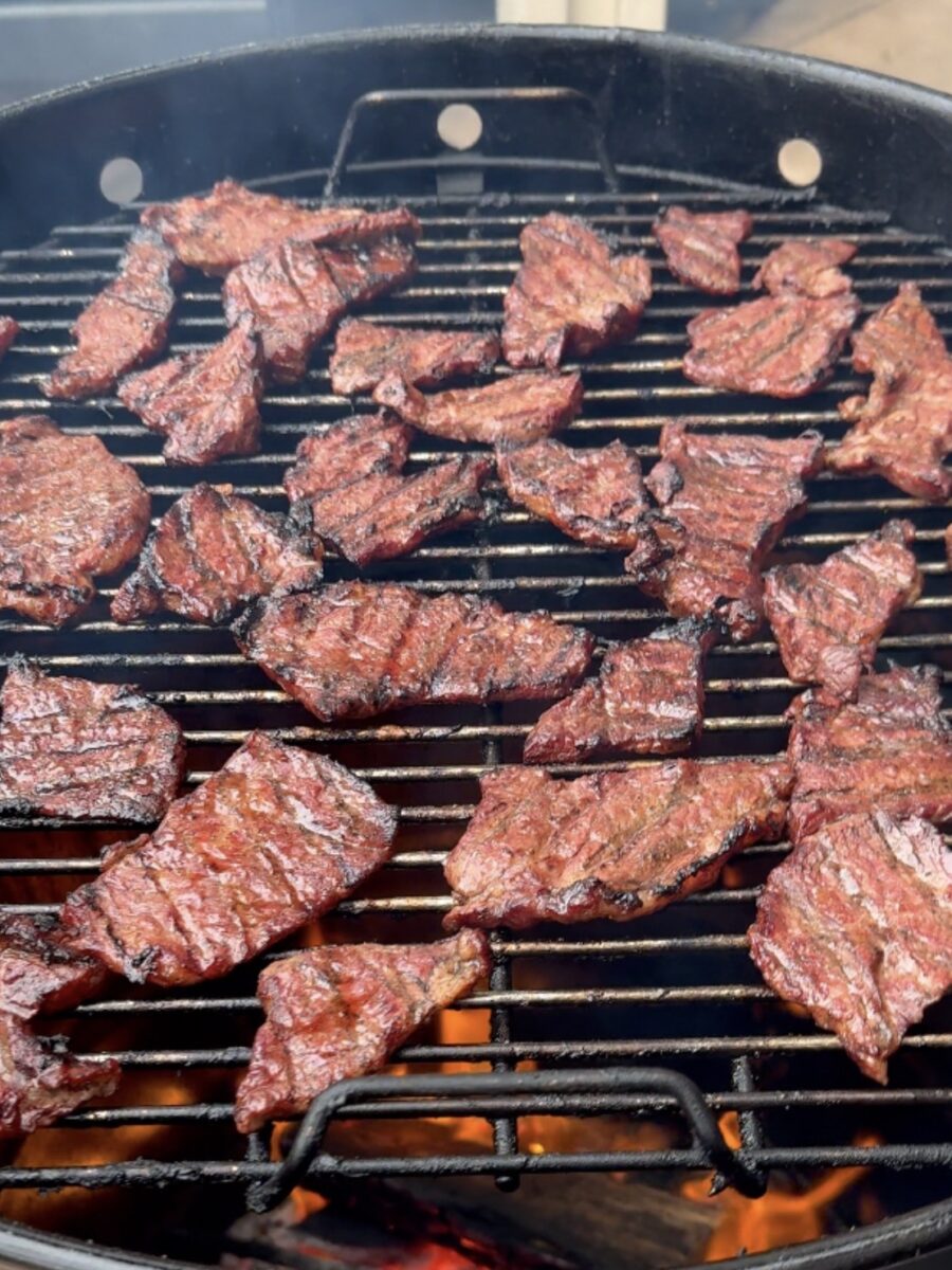 Grilled pieces of marinated meat cooking on a barbecue with flames visible beneath the metal grate.