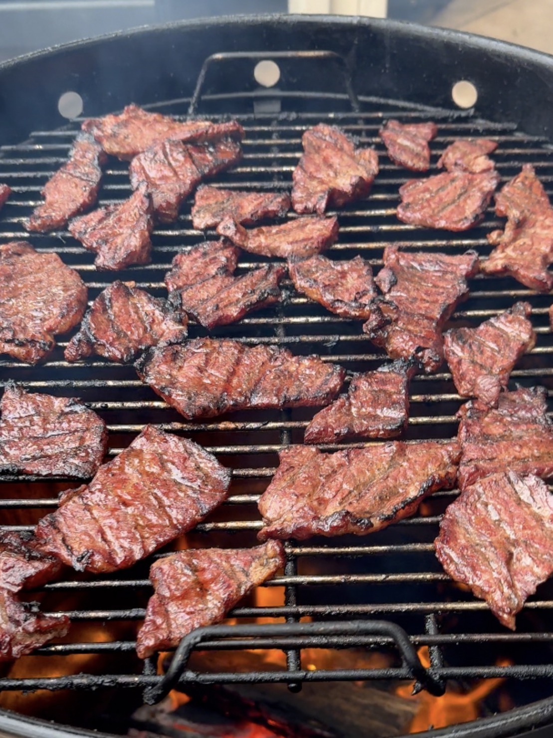 Grilled pieces of marinated meat cooking on a barbecue with flames visible beneath the metal grate.