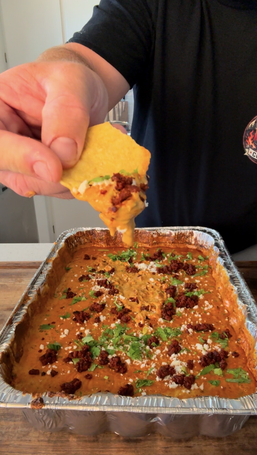 A hand holds a tortilla chip scooping cheesy, chili dip from a foil tray. The dip is topped with crumbled sausage, cheese, and chopped herbs.