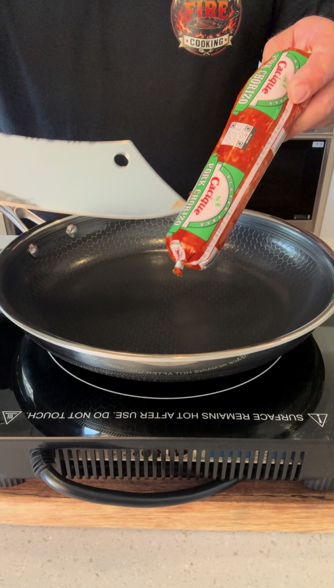A person holds a tube of Cacique chorizo sausage over a nonstick frying pan on an electric cooktop, preparing to cook.