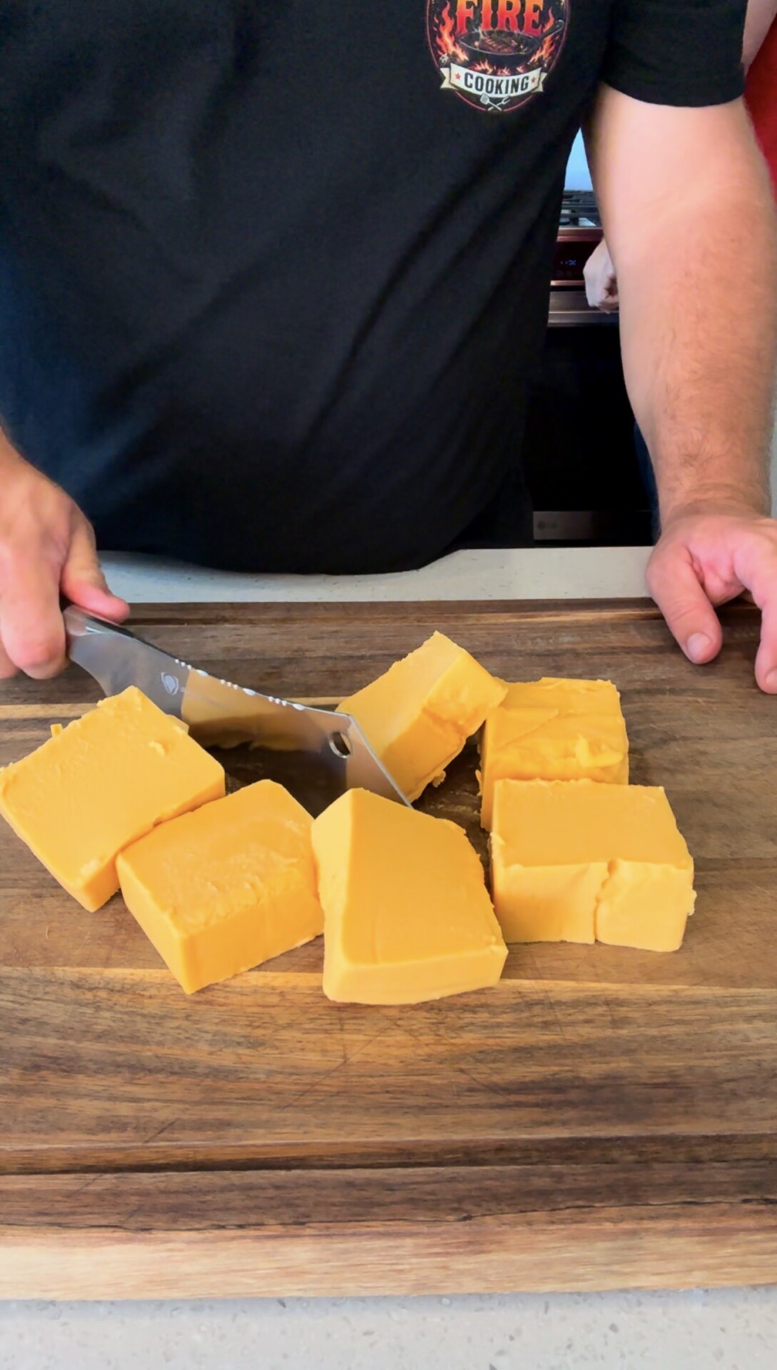 A person slices large blocks of orange cheddar cheese on a wooden cutting board with a large knife.
