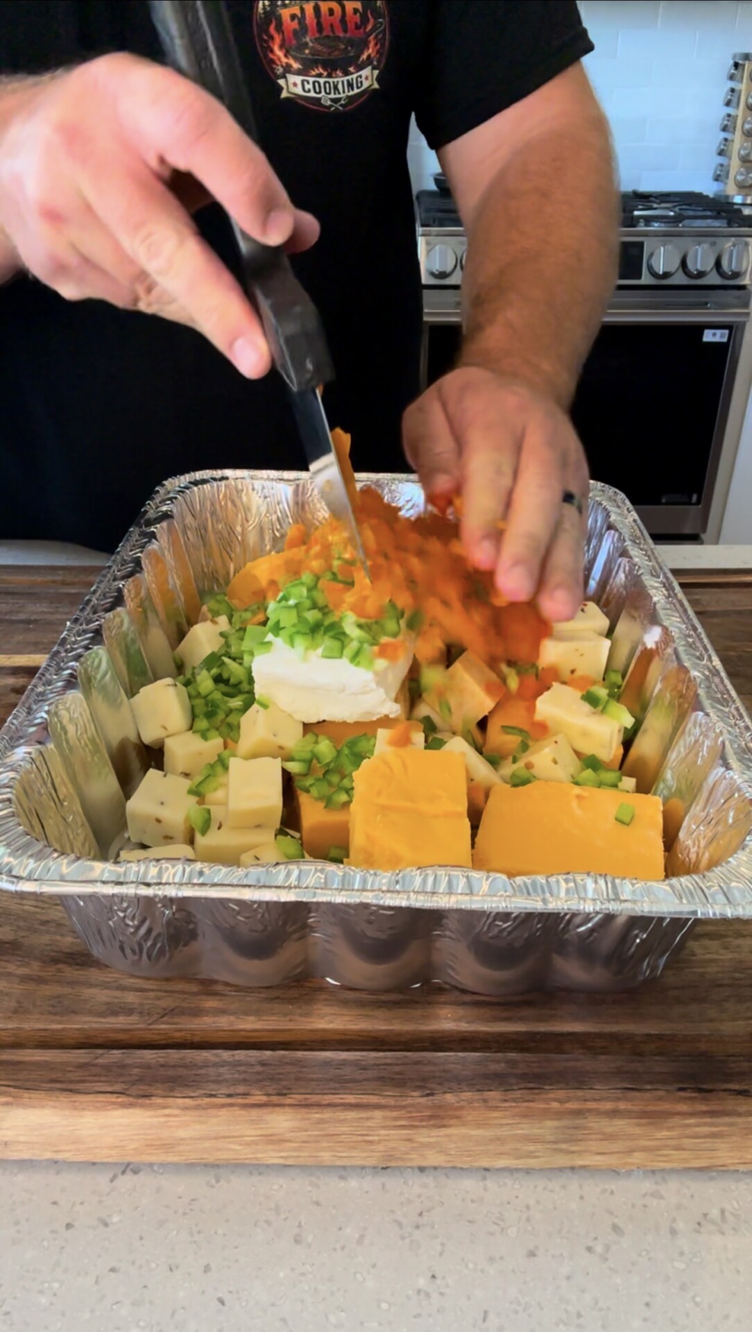 A person chops vegetables over a foil tray filled with cubed cheese, cream cheese, and chopped green onions, preparing ingredients on a wooden countertop in a kitchen.