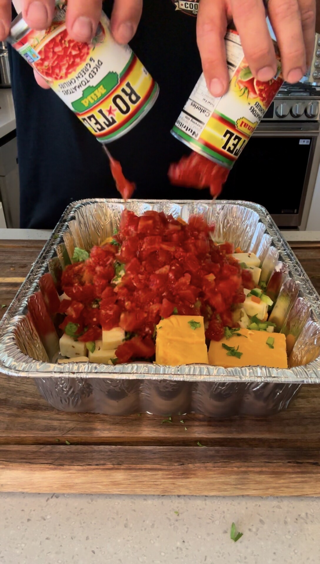 Two hands pour canned diced tomatoes onto a metal tray filled with cubed cheese, cream cheese, and chopped herbs, all sitting on a wooden countertop in a kitchen.