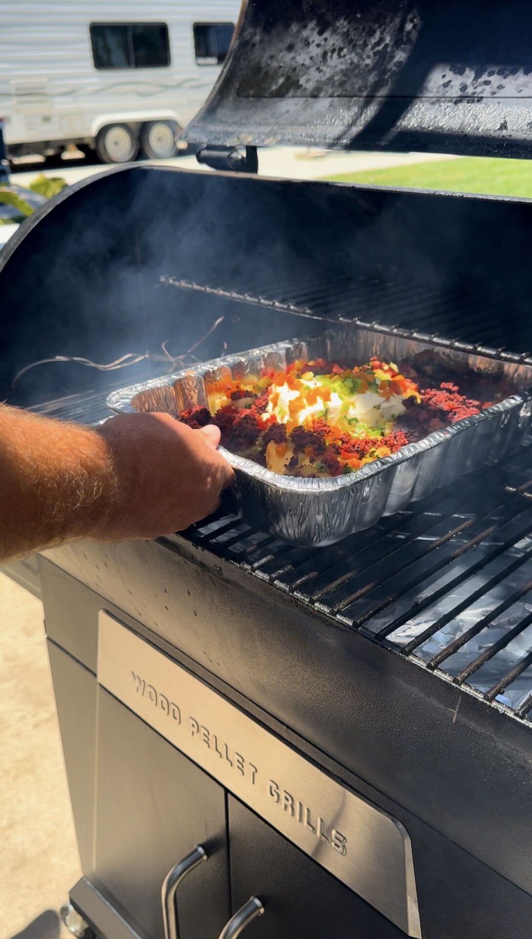 A person places a foil tray filled with colorful chopped veggies and ground meat onto a smoking grill labeled Wood Pellet Grills outside on a sunny day.