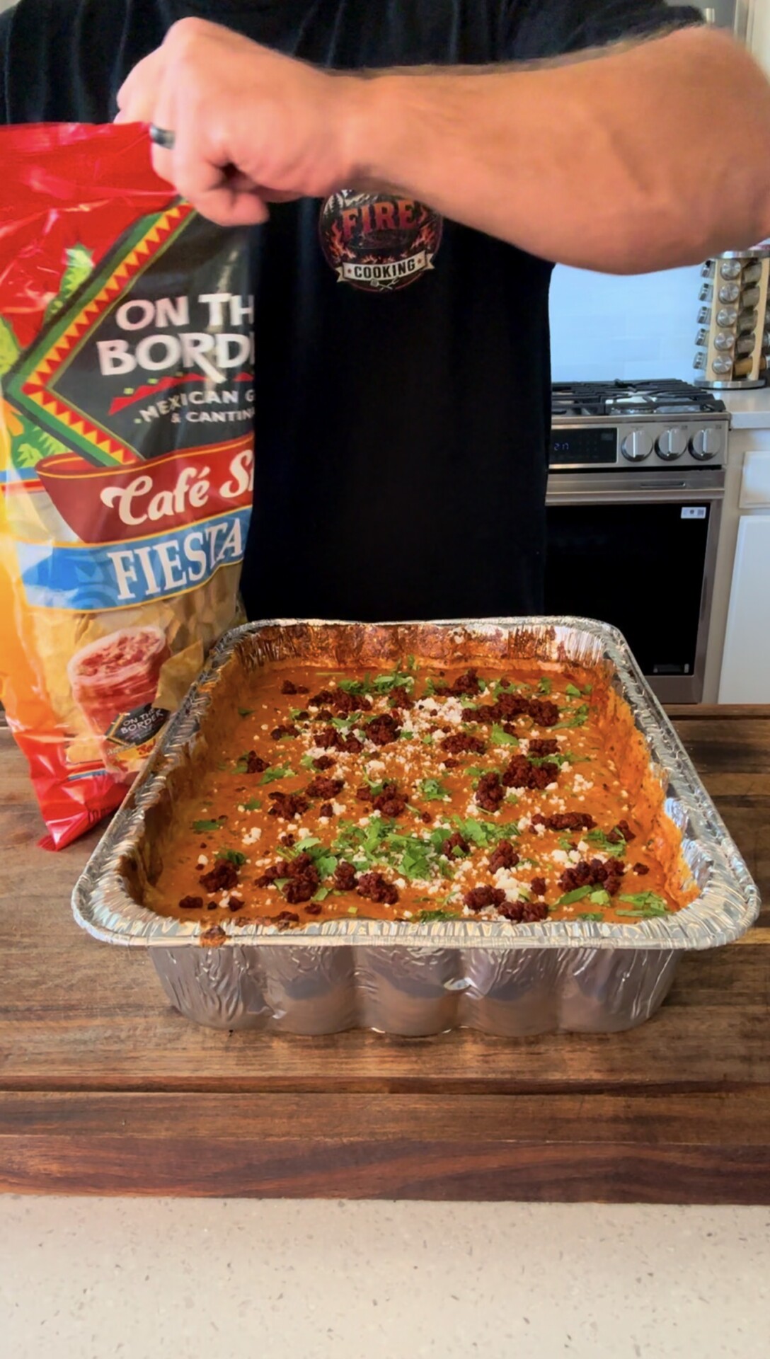 A person opens a bag of tortilla chips next to a large foil tray of cheesy dip topped with herbs and crumbled meat on a kitchen counter.