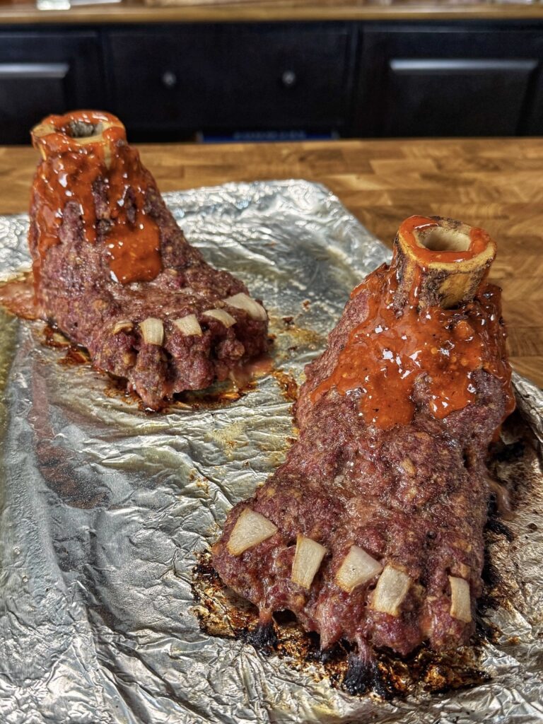 Two meatloaf sculptures shaped like feet, topped with bones for ankles and pieces of onion for toenails, sit on foil on a baking tray, covered in reddish sauce.