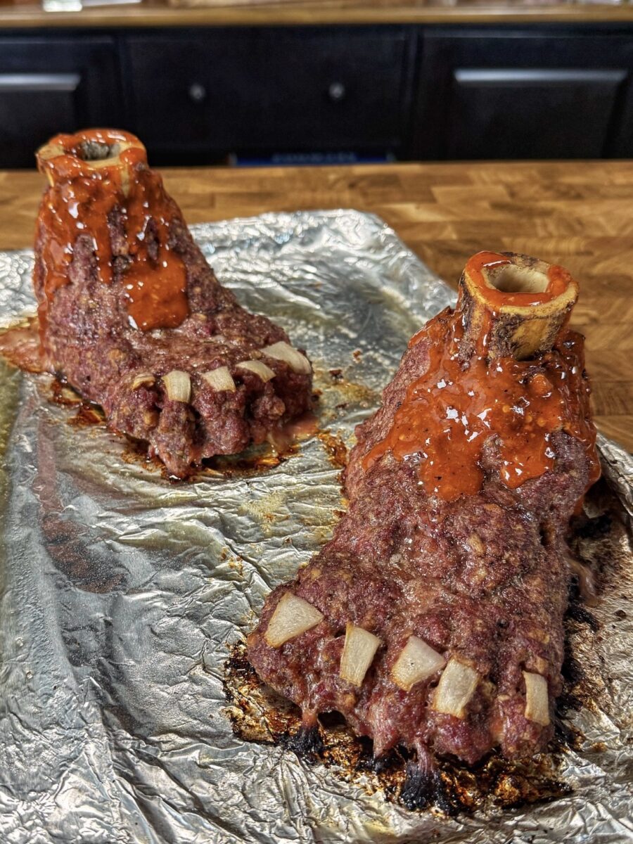 Two meatloaf sculptures shaped like feet, topped with bones for ankles and pieces of onion for toenails, sit on foil on a baking tray, covered in reddish sauce.