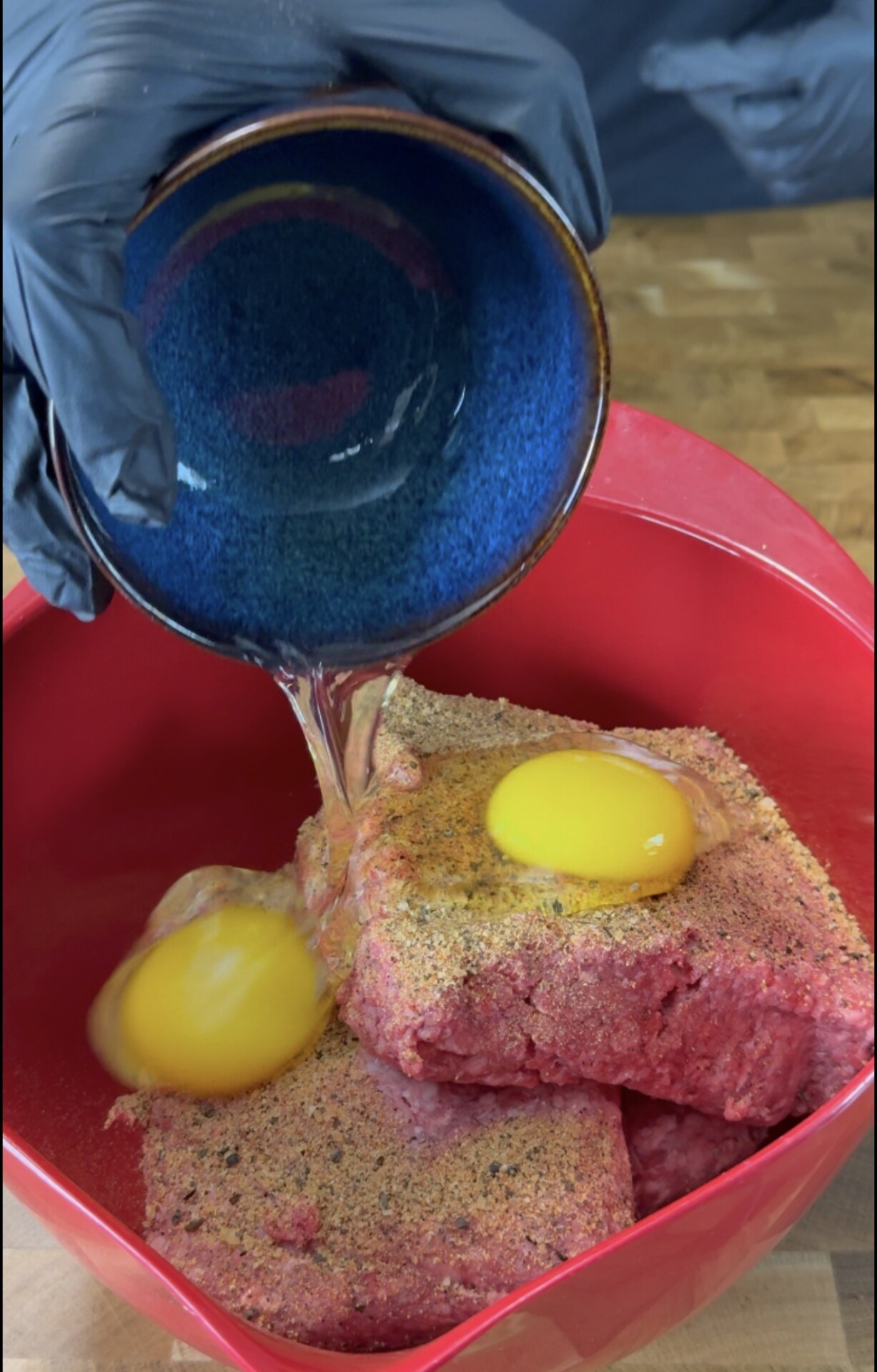 A gloved hand pours egg whites from a blue bowl onto ground beef and whole eggs in a red mixing bowl, preparing for a recipe.