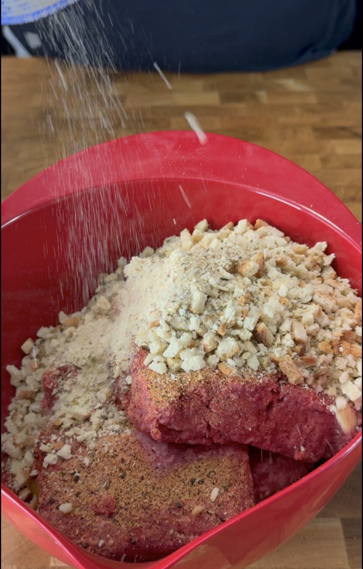 Seasonings and breadcrumbs are being sprinkled onto ground beef in a red bowl, placed on a wooden surface, preparing for mixing.