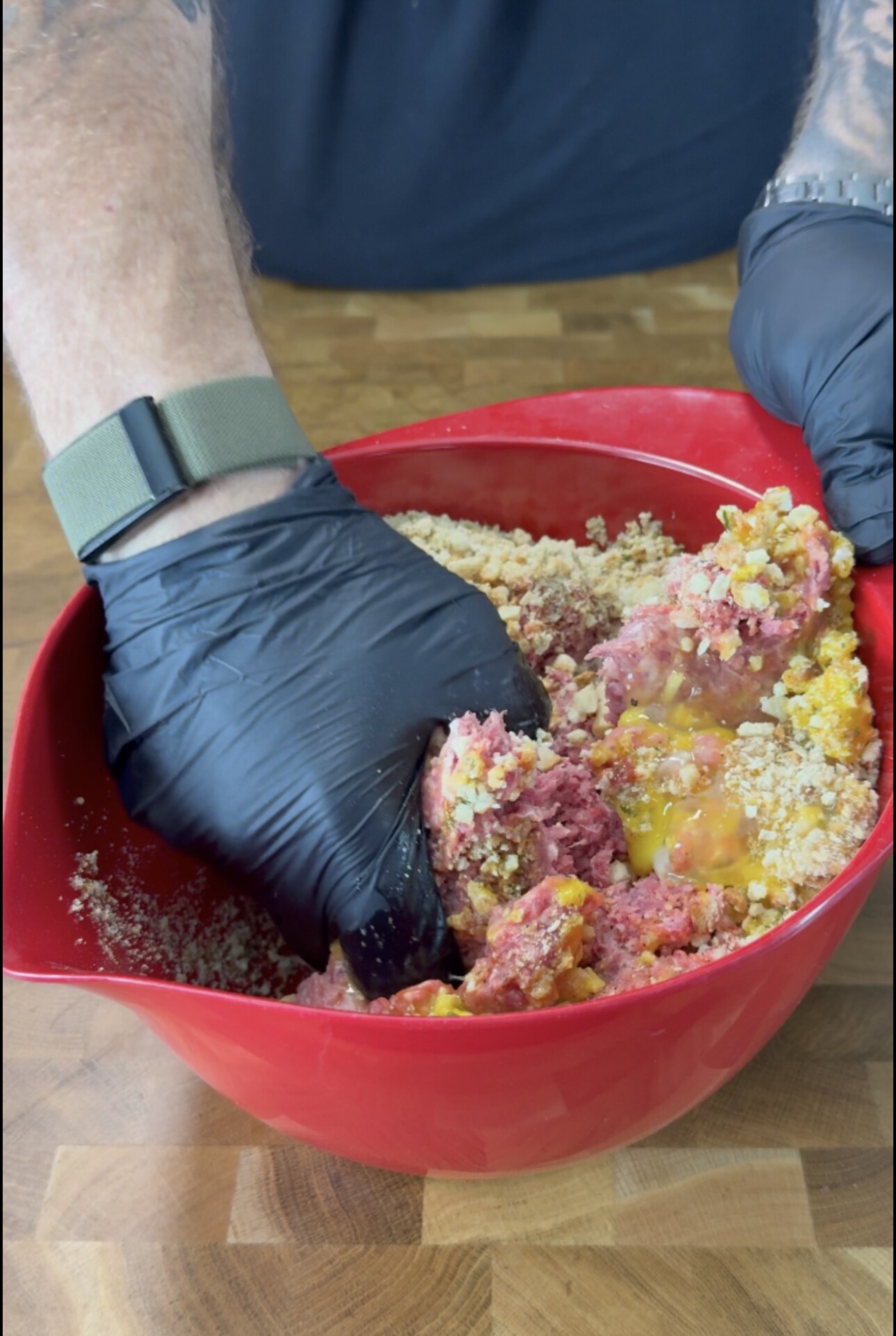 A person wearing black gloves mixes ground meat, eggs, breadcrumbs, and seasonings in a large red bowl on a wooden surface.