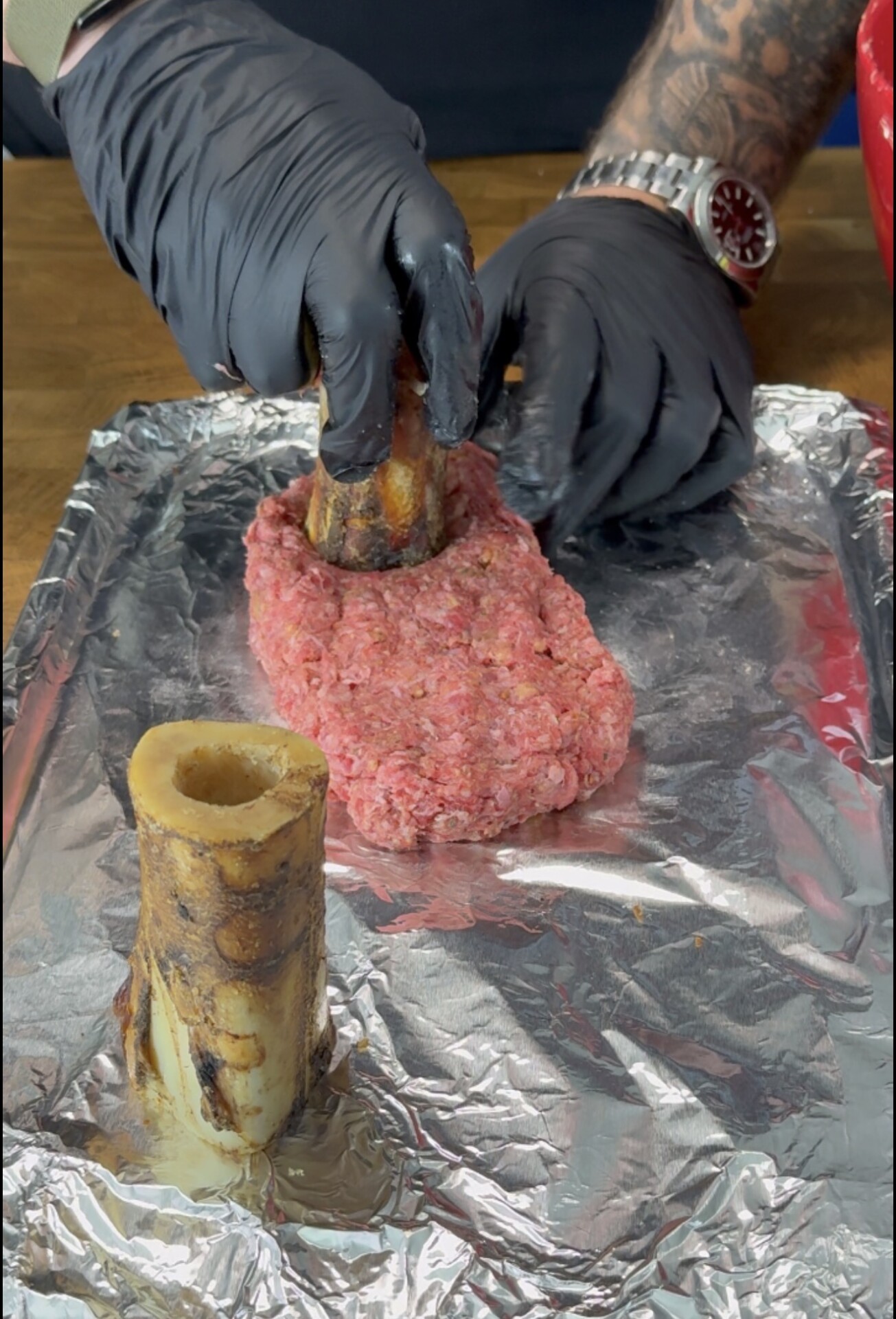A person wearing black gloves shapes raw ground beef around a bone marrow piece on a foil-lined tray, with another bone marrow segment nearby.