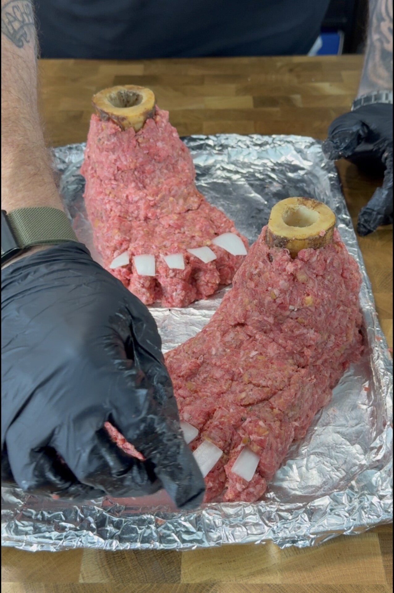 Two hands with black gloves shape ground meat into feet with bones as ankles and cubed onions as toenails, displayed on a foil-lined tray.