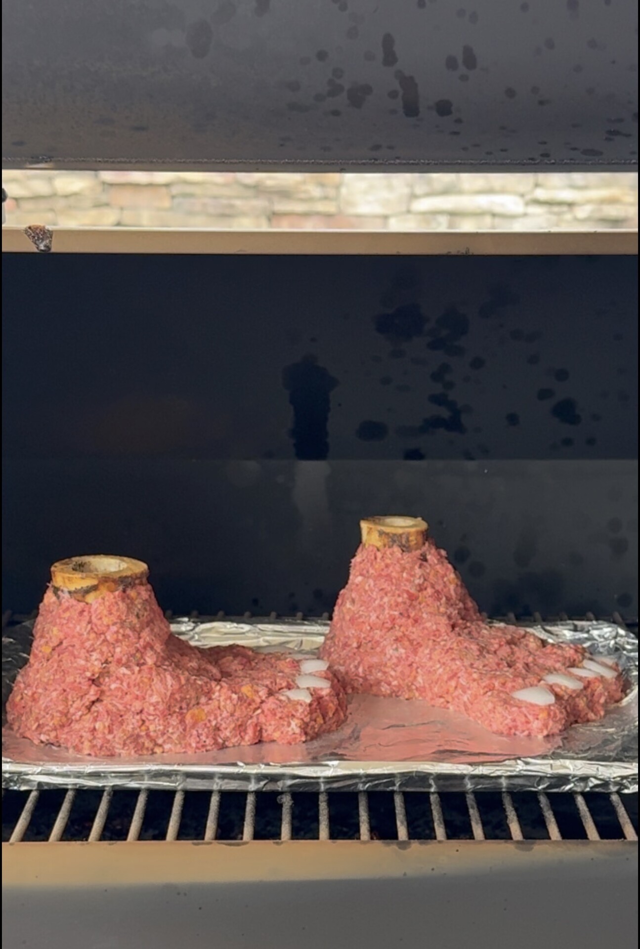 Two raw ground meat mixtures shaped like feet with bones at the ankles are placed on foil inside a grill or smoker, ready for cooking.