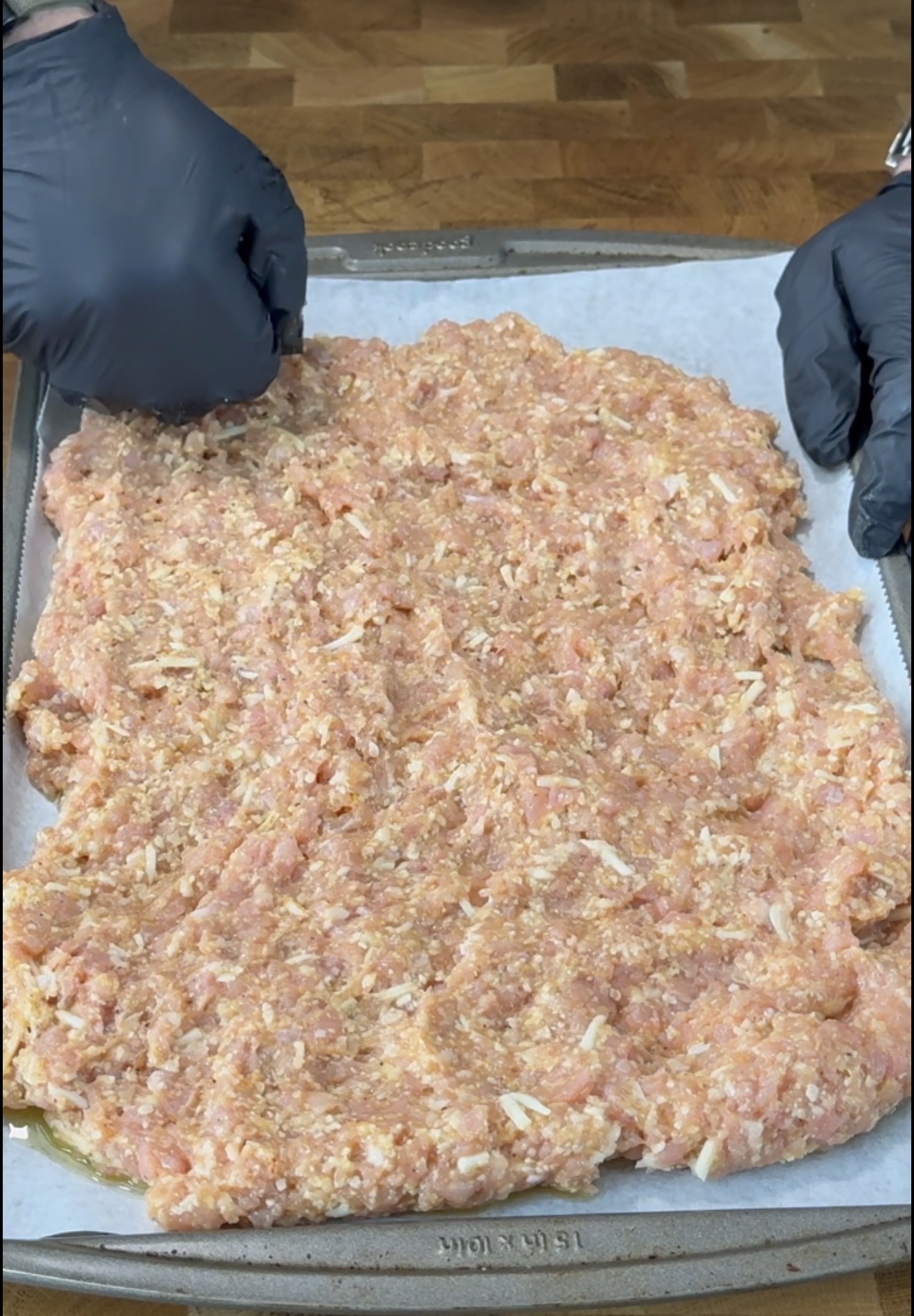 Ground meat mixture is being spread evenly on a parchment-lined baking sheet by two people wearing black gloves.