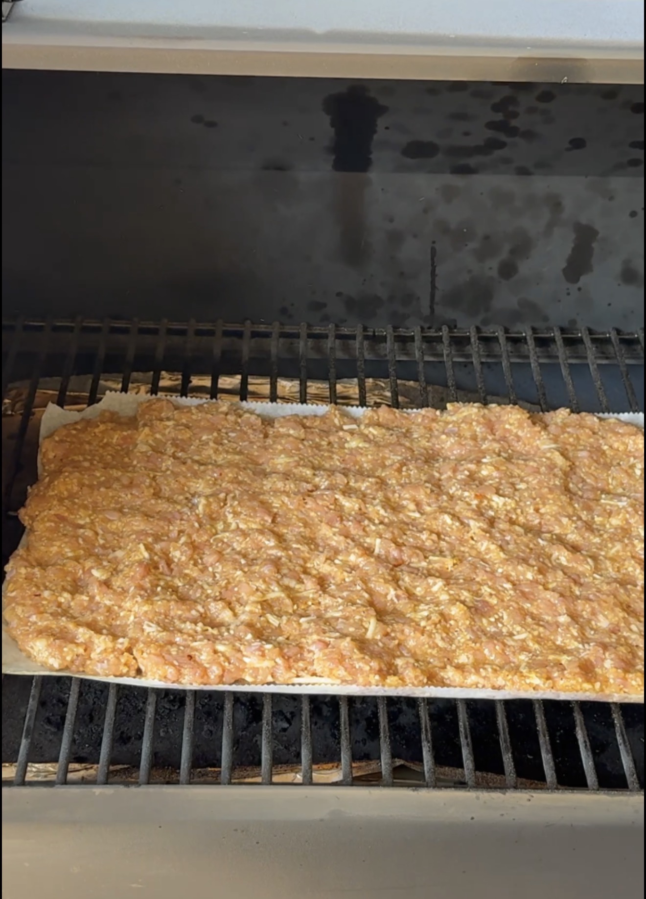 Raw ground meat mixture spread evenly on a rectangular tray inside a grill, with dark grill bars and a slightly stained metal surface visible in the background.