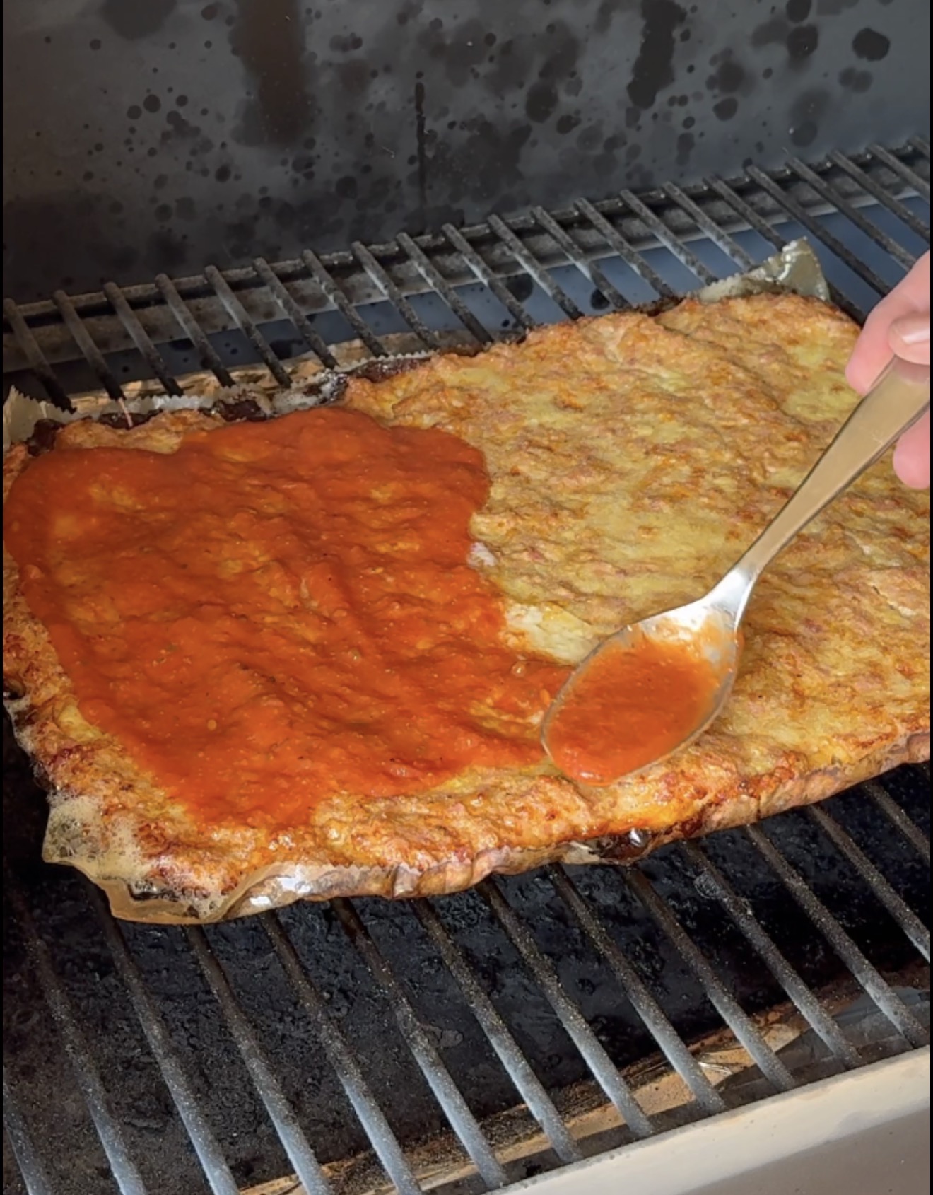 A hand spreads tomato sauce over a large rectangular pizza crust on a grill using a spoon. The crust is golden brown and only half is covered with sauce.