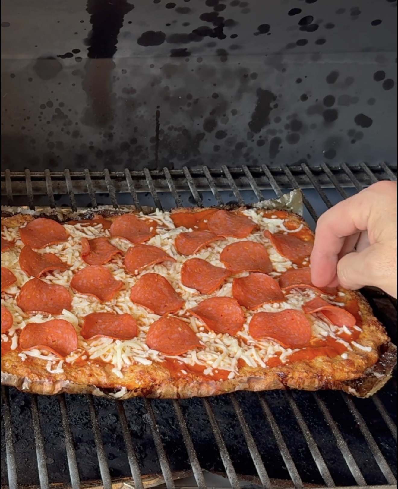 A hand is touching a rectangular pepperoni pizza with melted cheese on a grill grate, with dark stains visible on the grills interior.