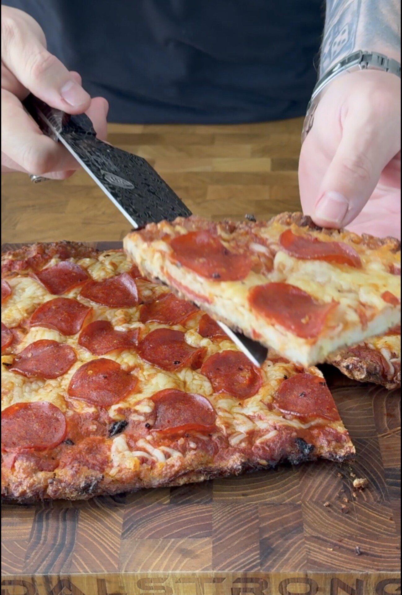 A person lifts a rectangular slice of pepperoni pizza from a wooden cutting board, revealing melted cheese and crispy edges.