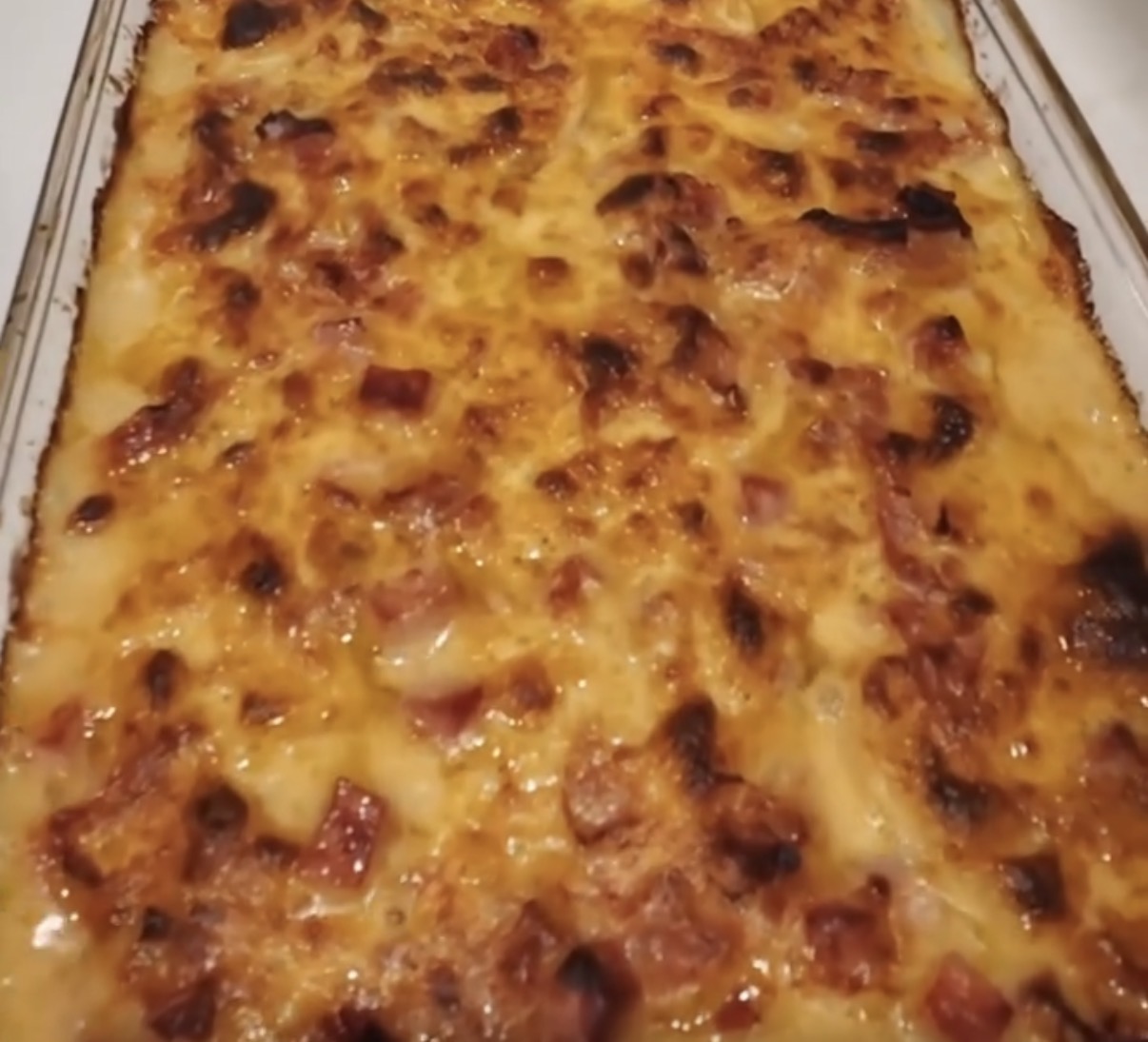 A close-up of a golden-brown baked casserole in a glass dish, with visible browned cheese and diced pieces of meat throughout the surface.