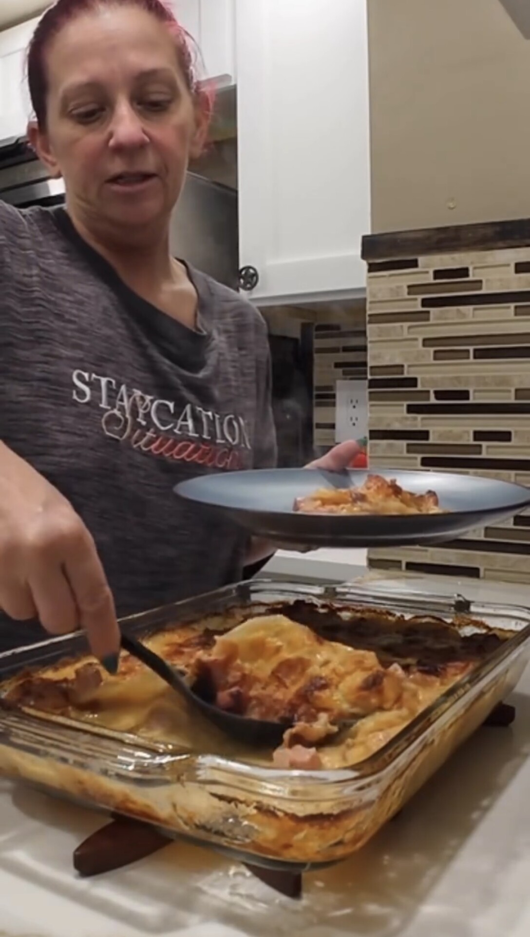 A person serves a portion of baked lasagna from a glass dish onto a blue plate in a kitchen with beige and brown tiled backsplash.