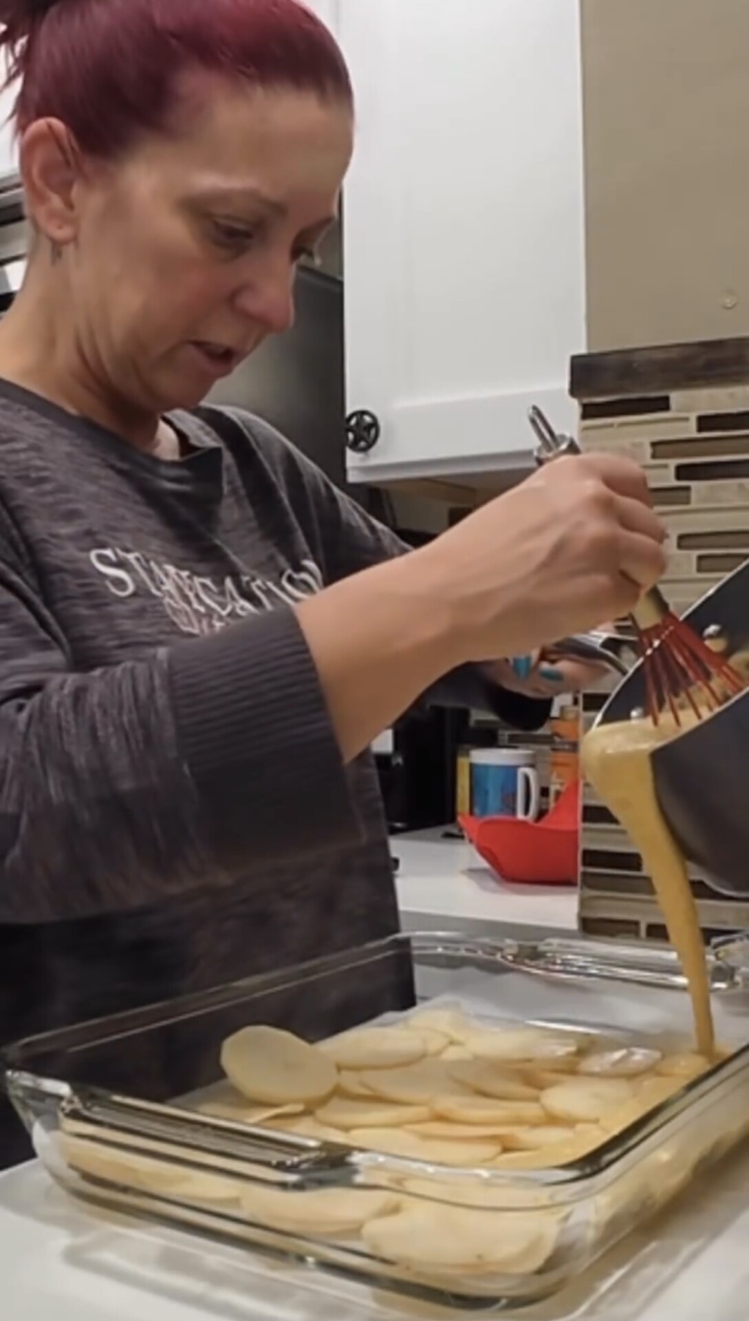 A woman with red hair pours a yellow sauce over sliced potatoes in a glass baking dish in a kitchen.