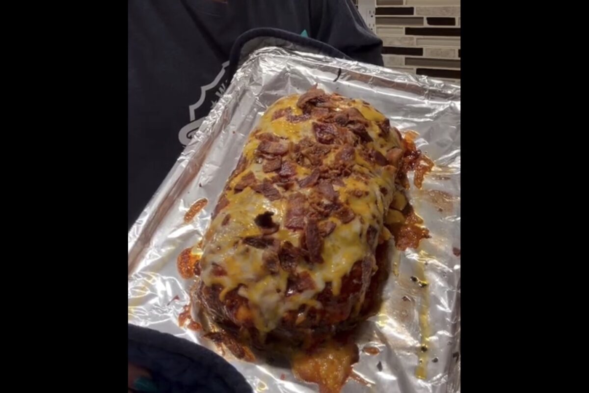 A person holds a tray with a freshly baked meatloaf topped with melted cheese and bacon bits, sitting on aluminum foil in a kitchen.