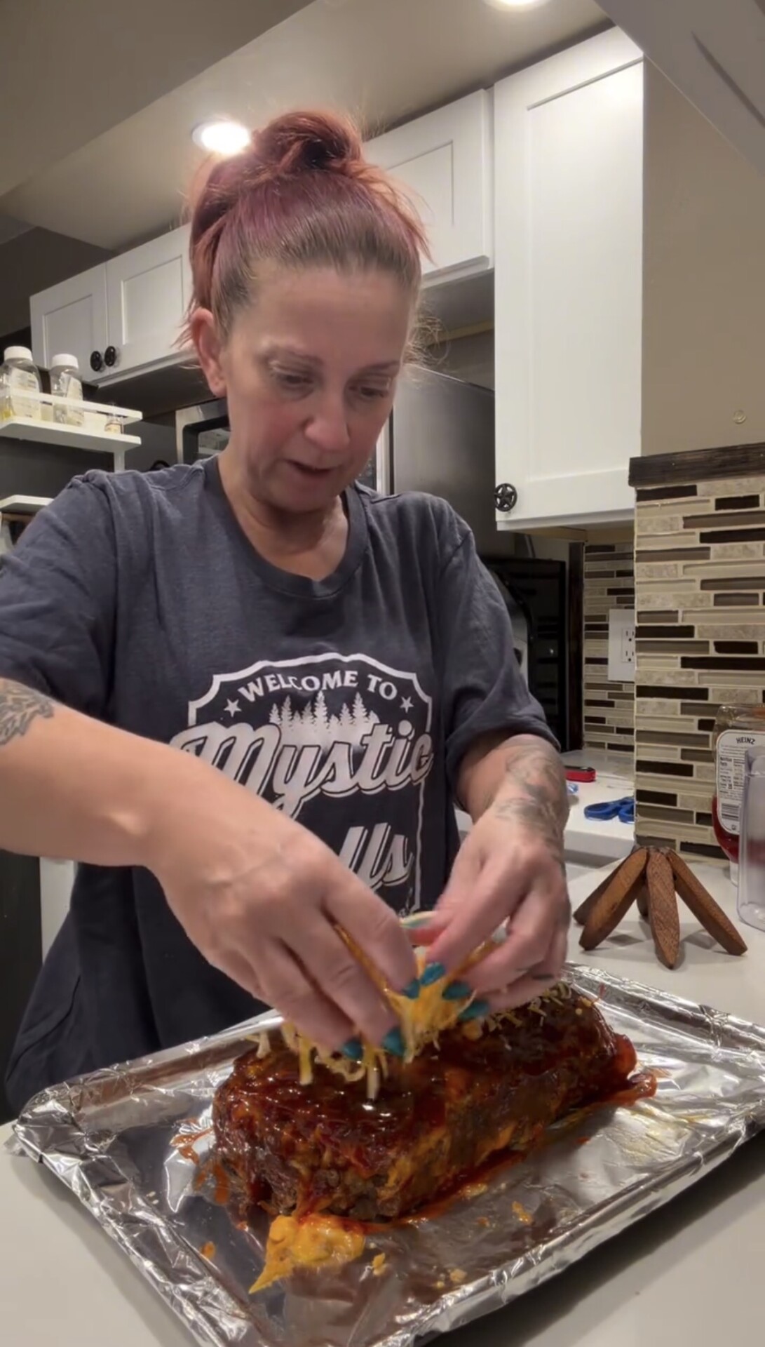 A woman with reddish hair in a ponytail adds toppings to a meatloaf on a foil-lined tray in a kitchen, wearing a Welcome to Mystic t-shirt.