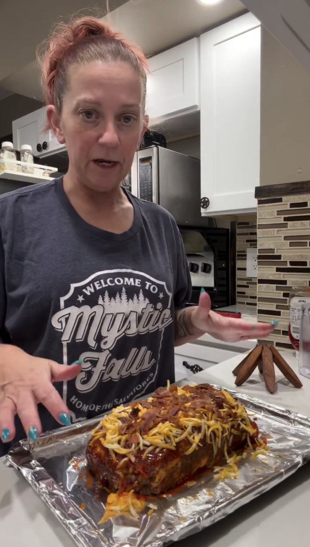A woman stands in a kitchen with her hands near a foil-covered tray holding a cheese-topped meatloaf. She wears a Mystic Falls T-shirt and looks toward the camera.