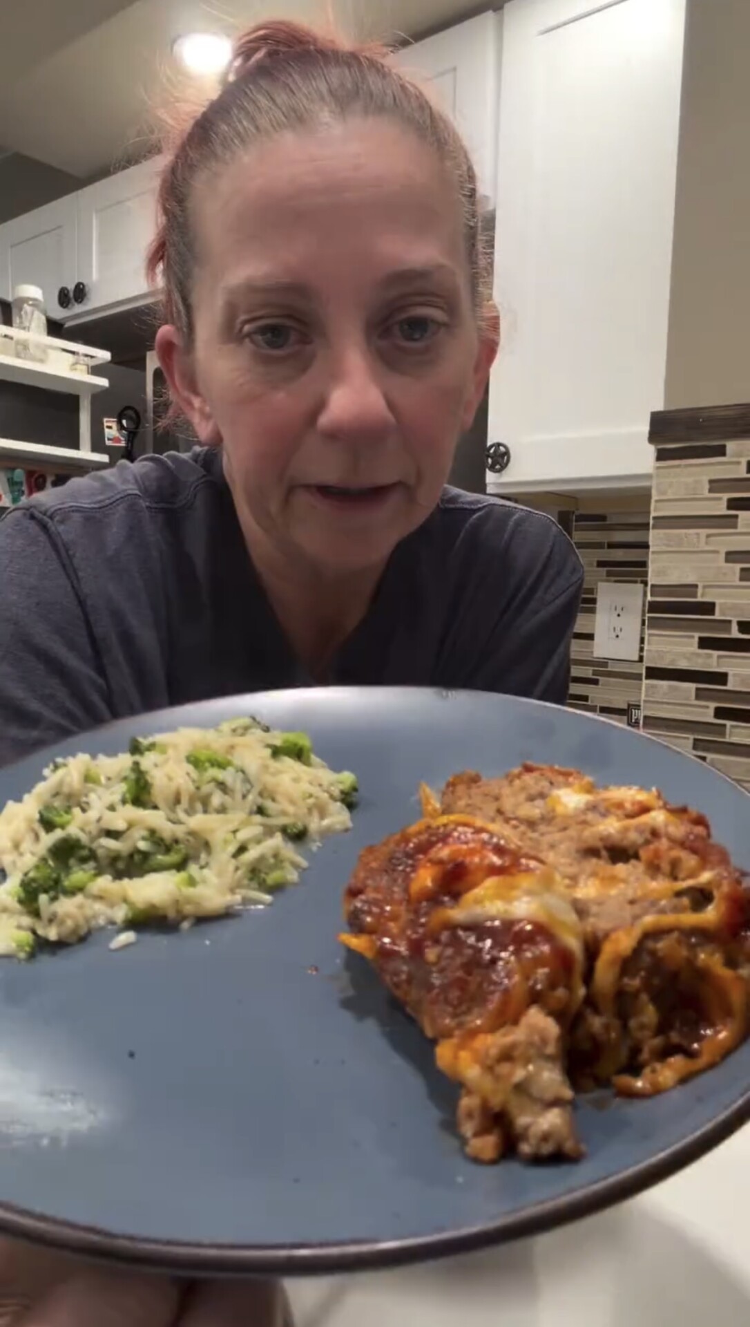 A woman holds a plate with a serving of lasagna and a side of rice mixed with green vegetables, standing in a kitchen with tiled backsplash.