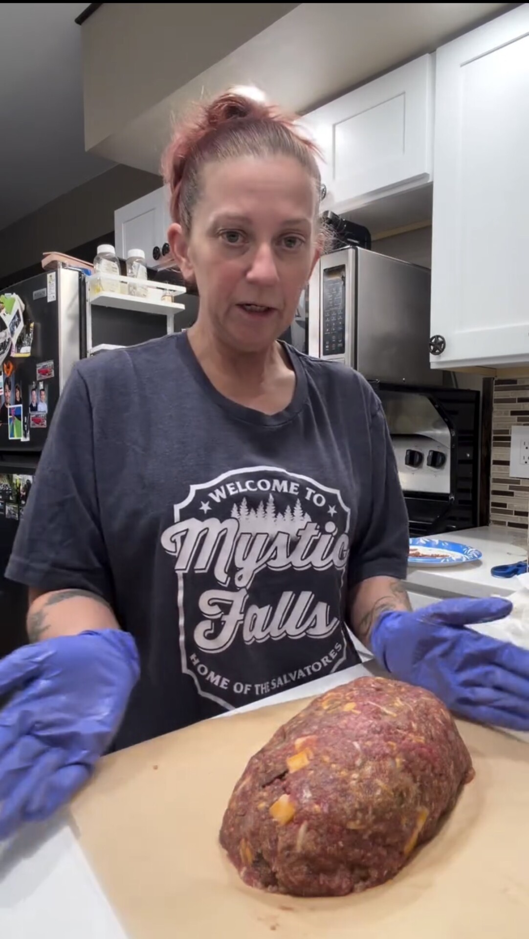 A person wearing blue gloves and a Mystic Falls t-shirt stands in a kitchen, presenting a large, uncooked meatloaf on parchment paper.