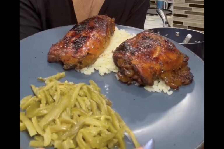 A blue plate holds two pieces of glazed, grilled chicken on a bed of white rice, with a serving of cooked green beans on the side. A person in dark clothing holds the plate in a kitchen setting.