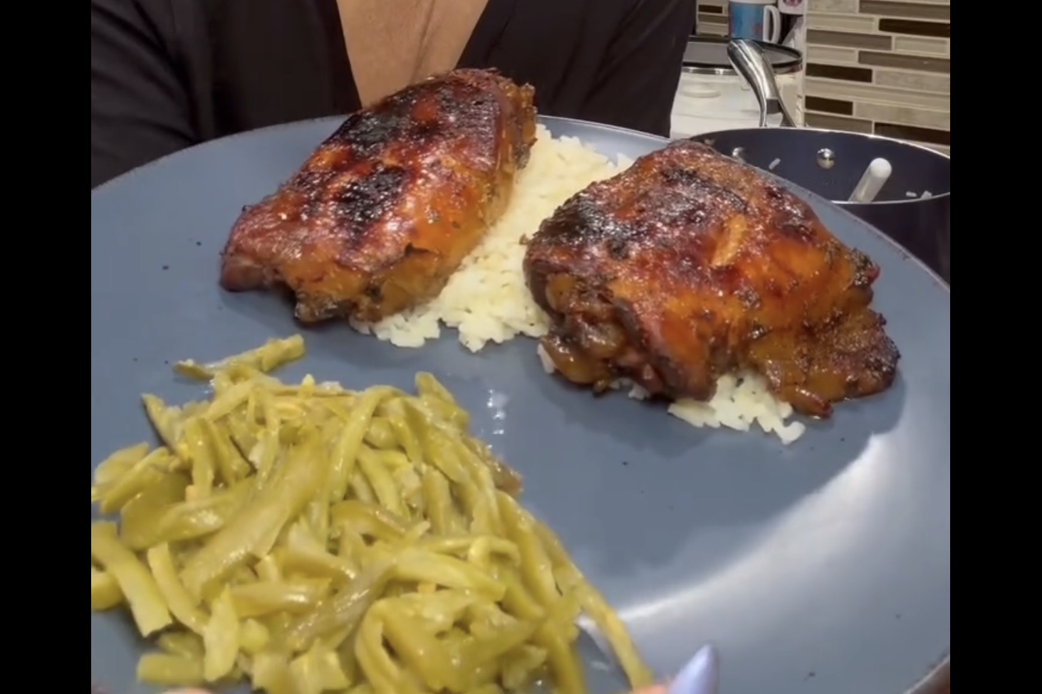 A blue plate holds two pieces of glazed, grilled chicken on a bed of white rice, with a serving of cooked green beans on the side. A person in dark clothing holds the plate in a kitchen setting.
