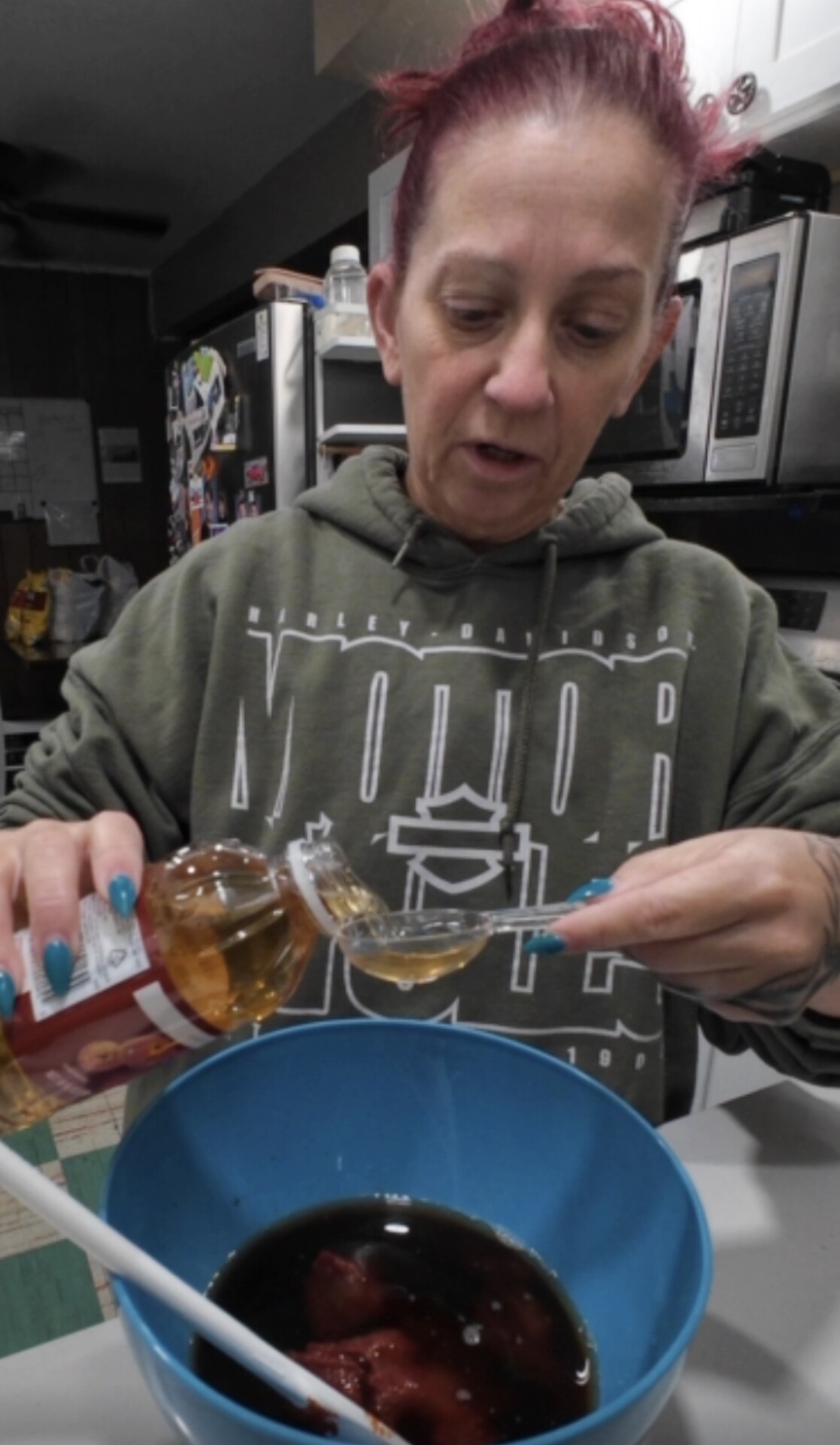 A person with red hair pours liquid from a bottle into a spoon over a blue bowl filled with dark-colored ingredients in a kitchen.