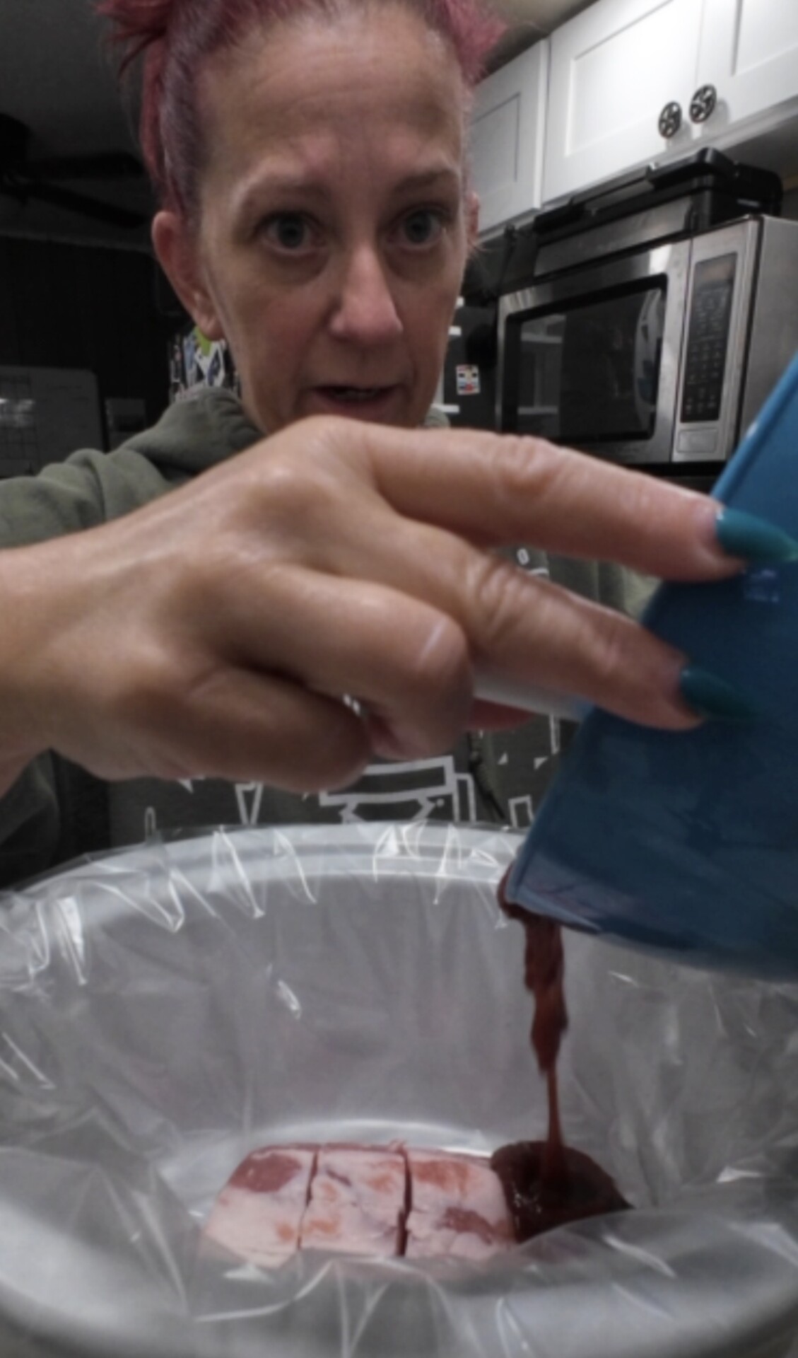 A person pours a liquid from a blue container into a slow cooker lined with plastic, with raw meat inside. Kitchen appliances are visible in the background.