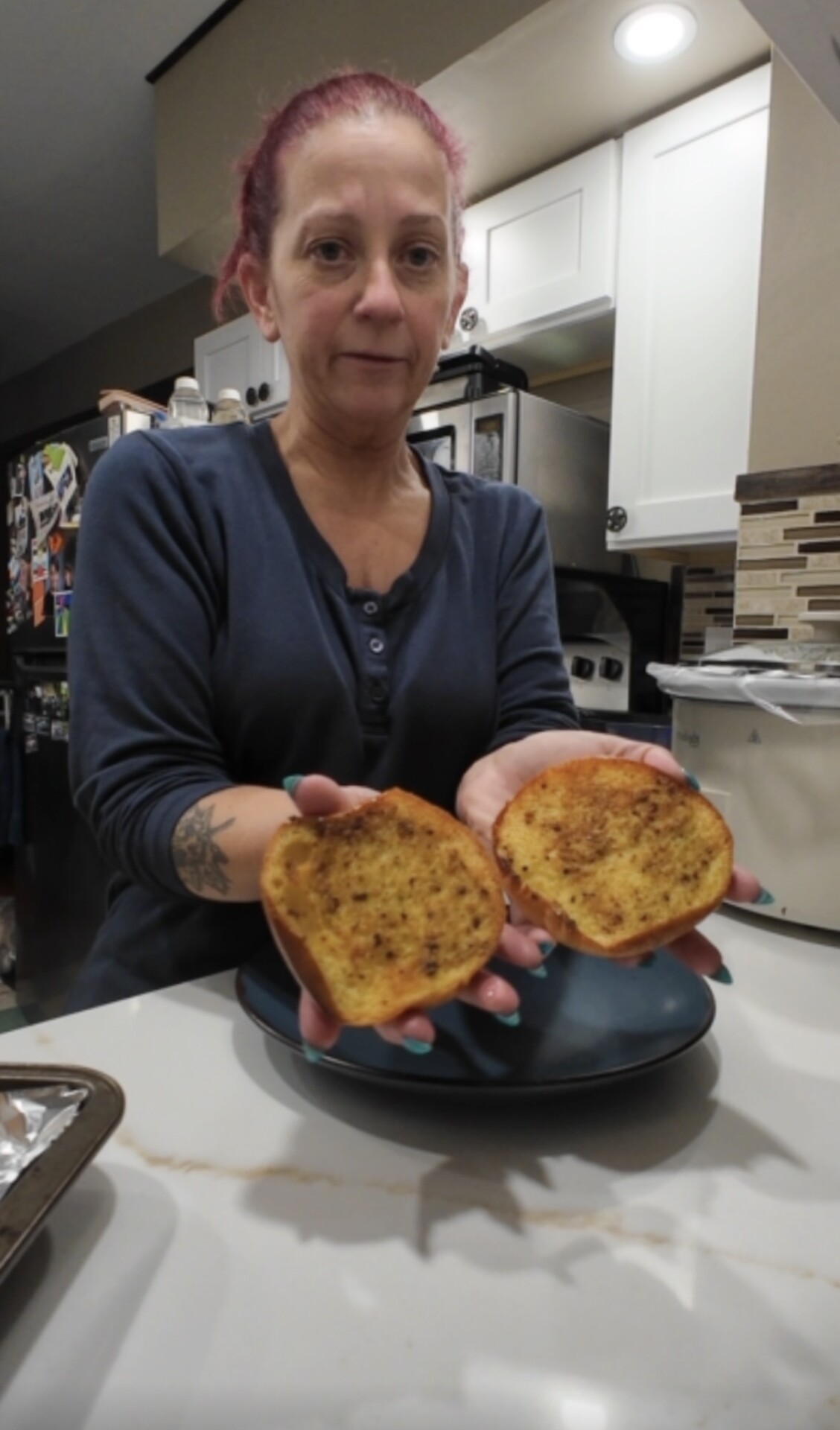 A woman with red hair in a blue shirt holds two halves of toasted, seasoned bread over a kitchen counter, facing the camera.