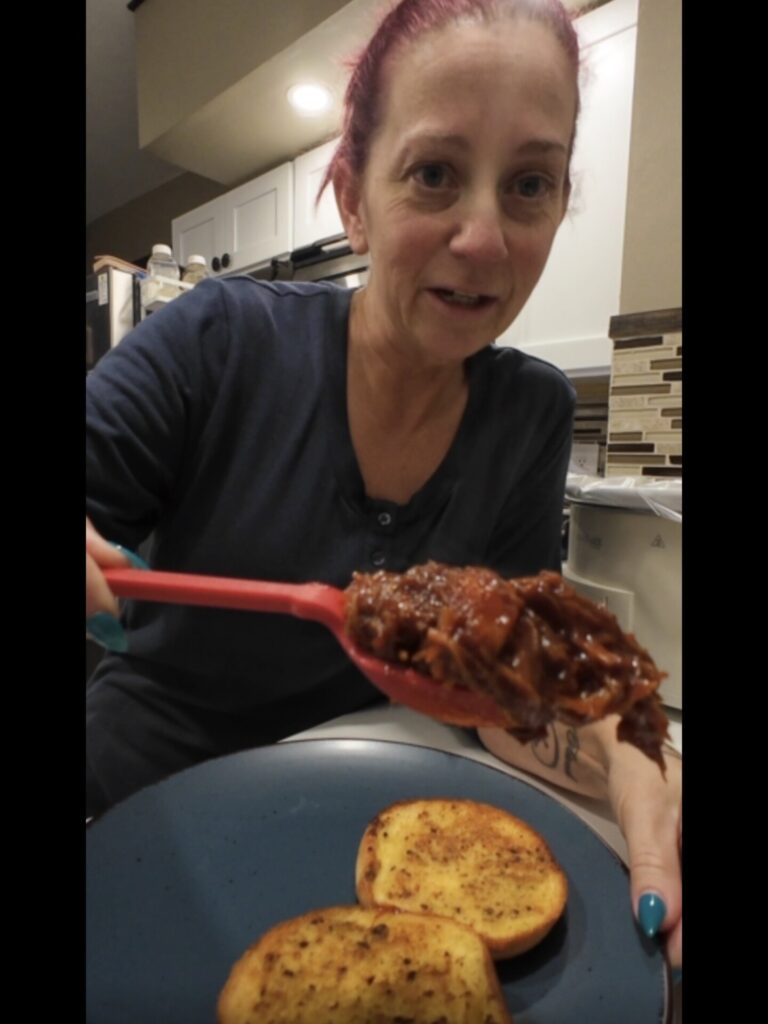 A woman with red hair holds a spoonful of shredded meat over a blue plate with two slices of toasted bread in a kitchen.
