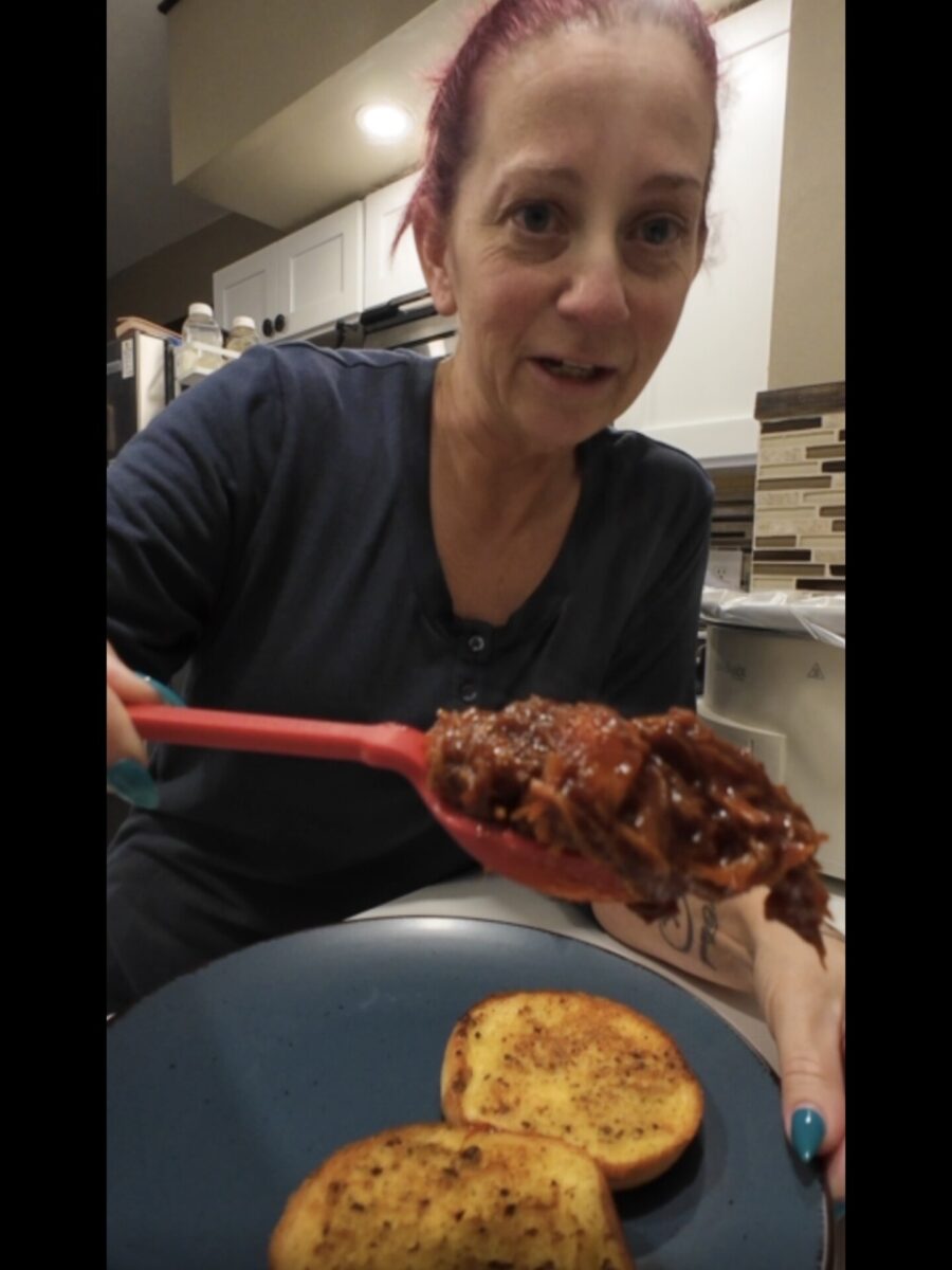A woman with red hair holds a spoonful of shredded meat over a blue plate with two slices of toasted bread in a kitchen.