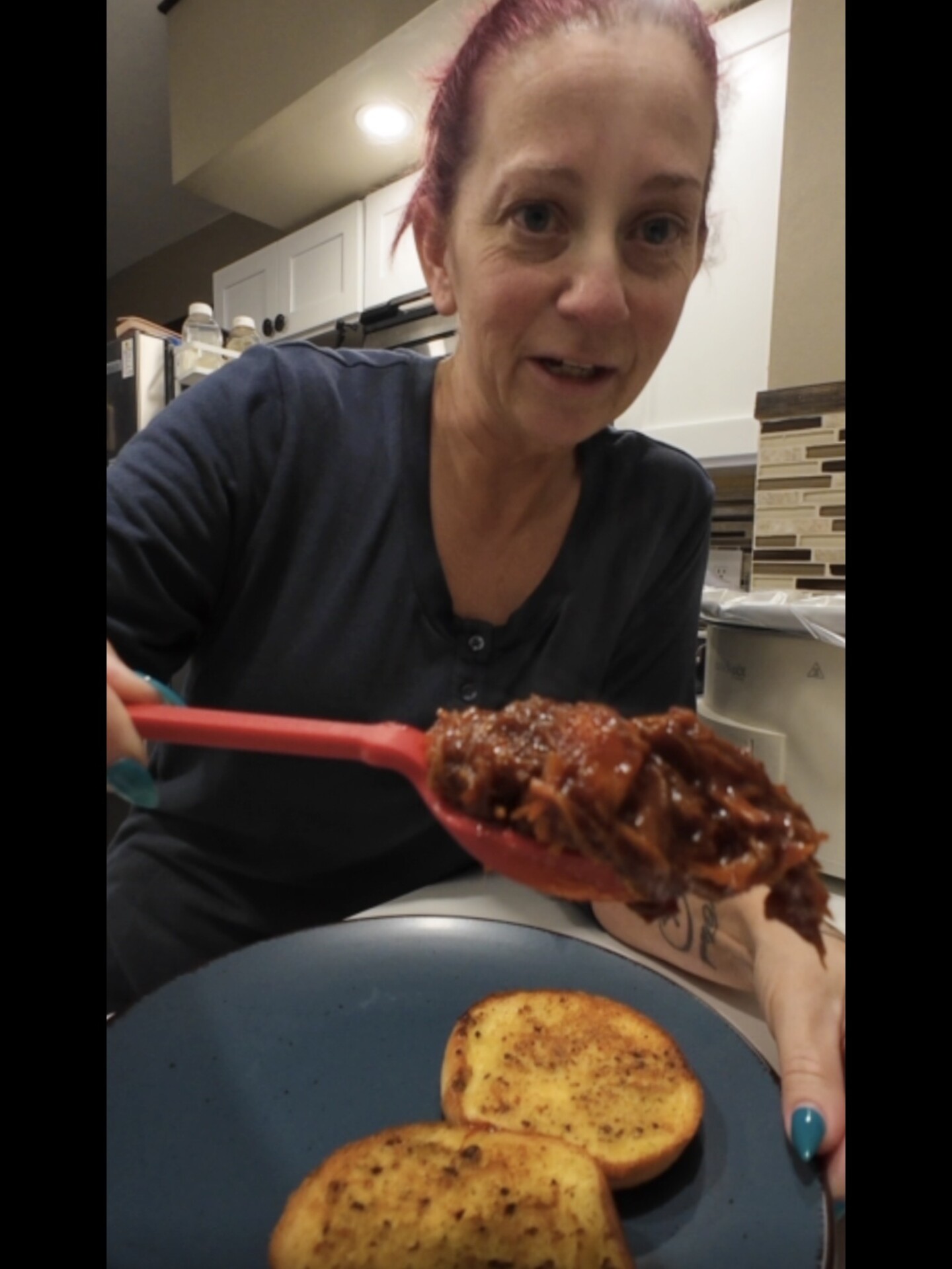 A woman with red hair holds a spoonful of shredded meat over a blue plate with two slices of toasted bread in a kitchen.