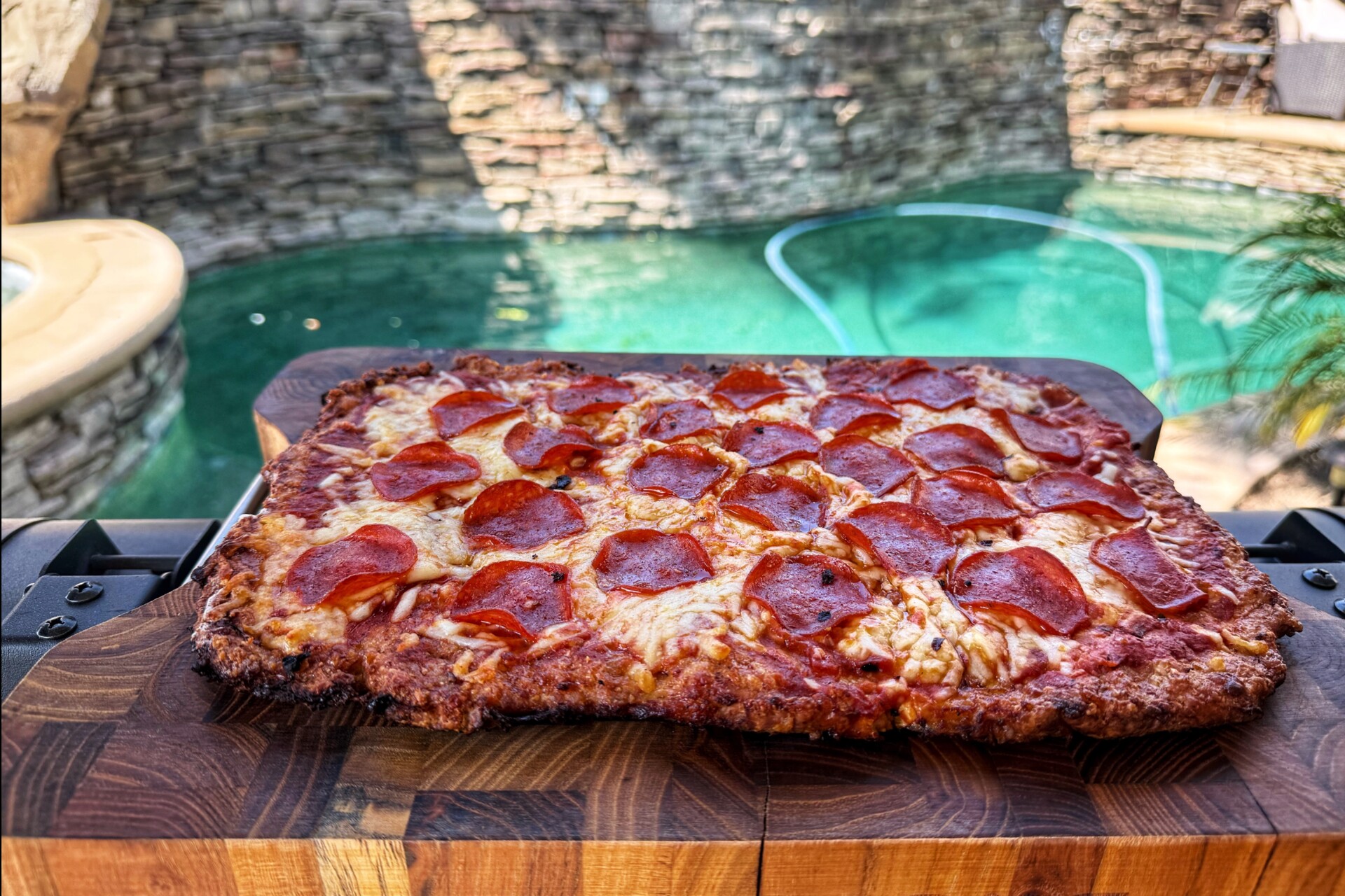 A rectangular pepperoni pizza on a wooden cutting board is in focus, with a clear swimming pool and stone wall in the sunny background.