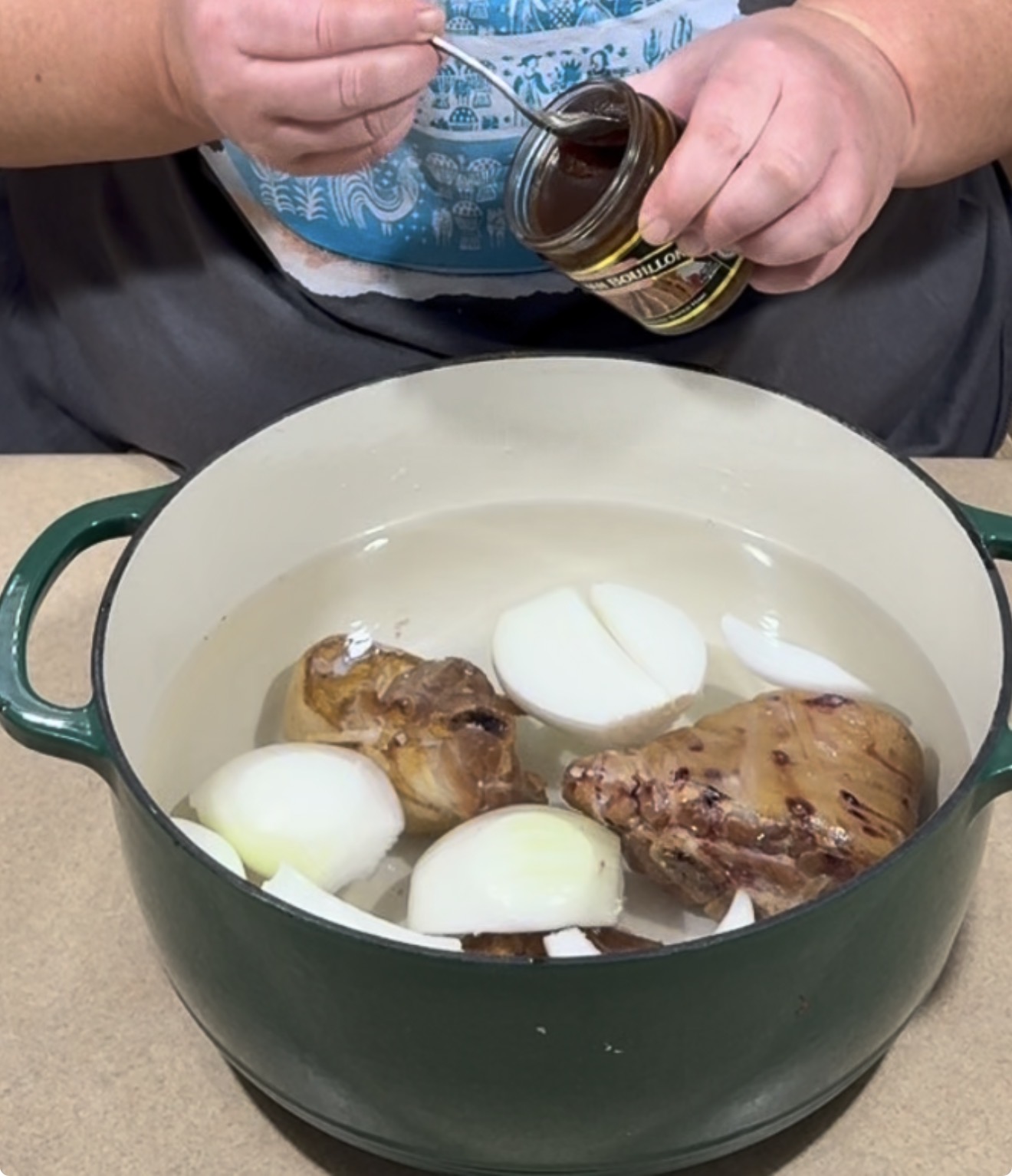 A person adds beef base from a jar into a green Dutch oven filled with water, onion halves, and meat pieces.