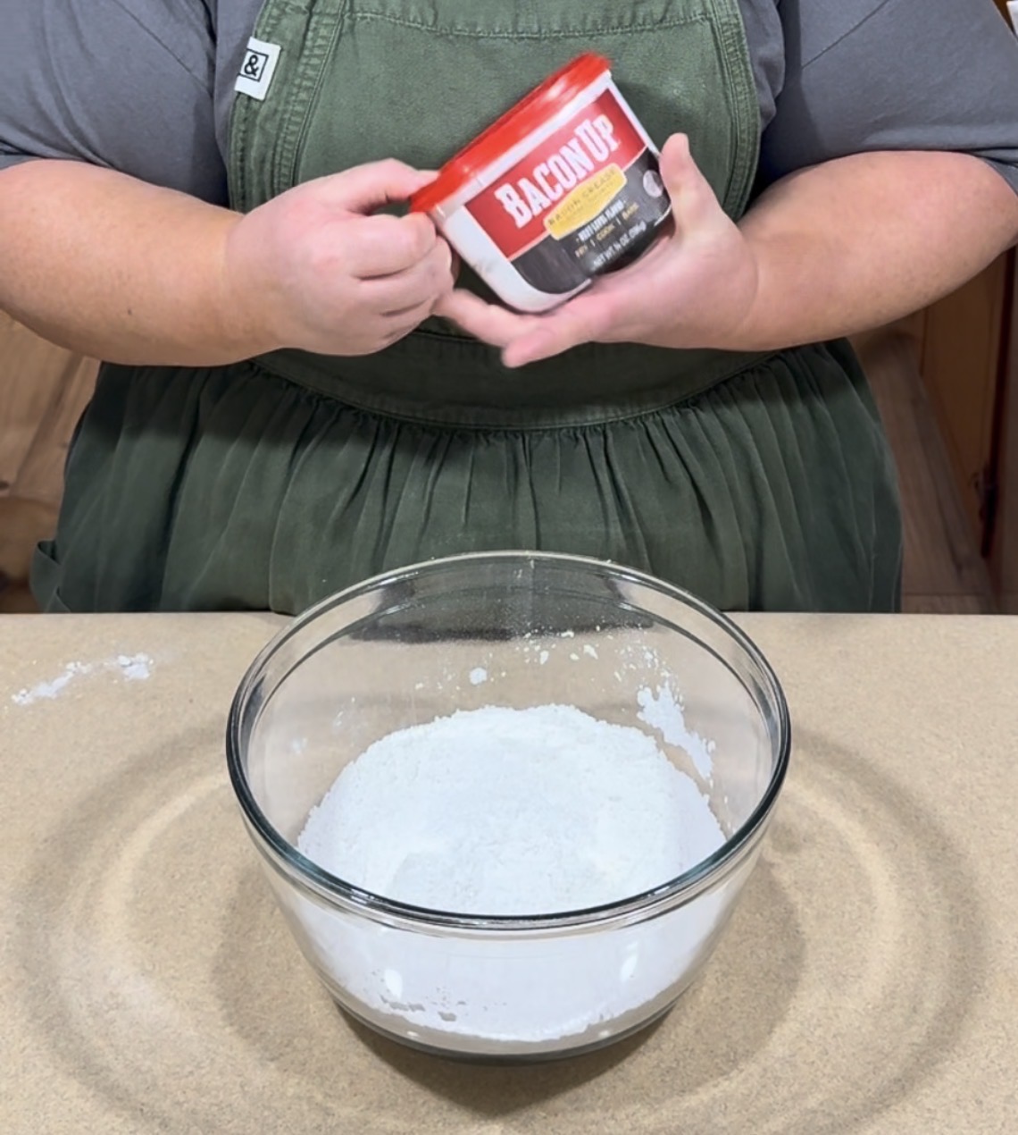 A person in a green apron holds a red tub labeled Bacon Up over a glass bowl filled with white powder, likely flour, on a kitchen counter.
