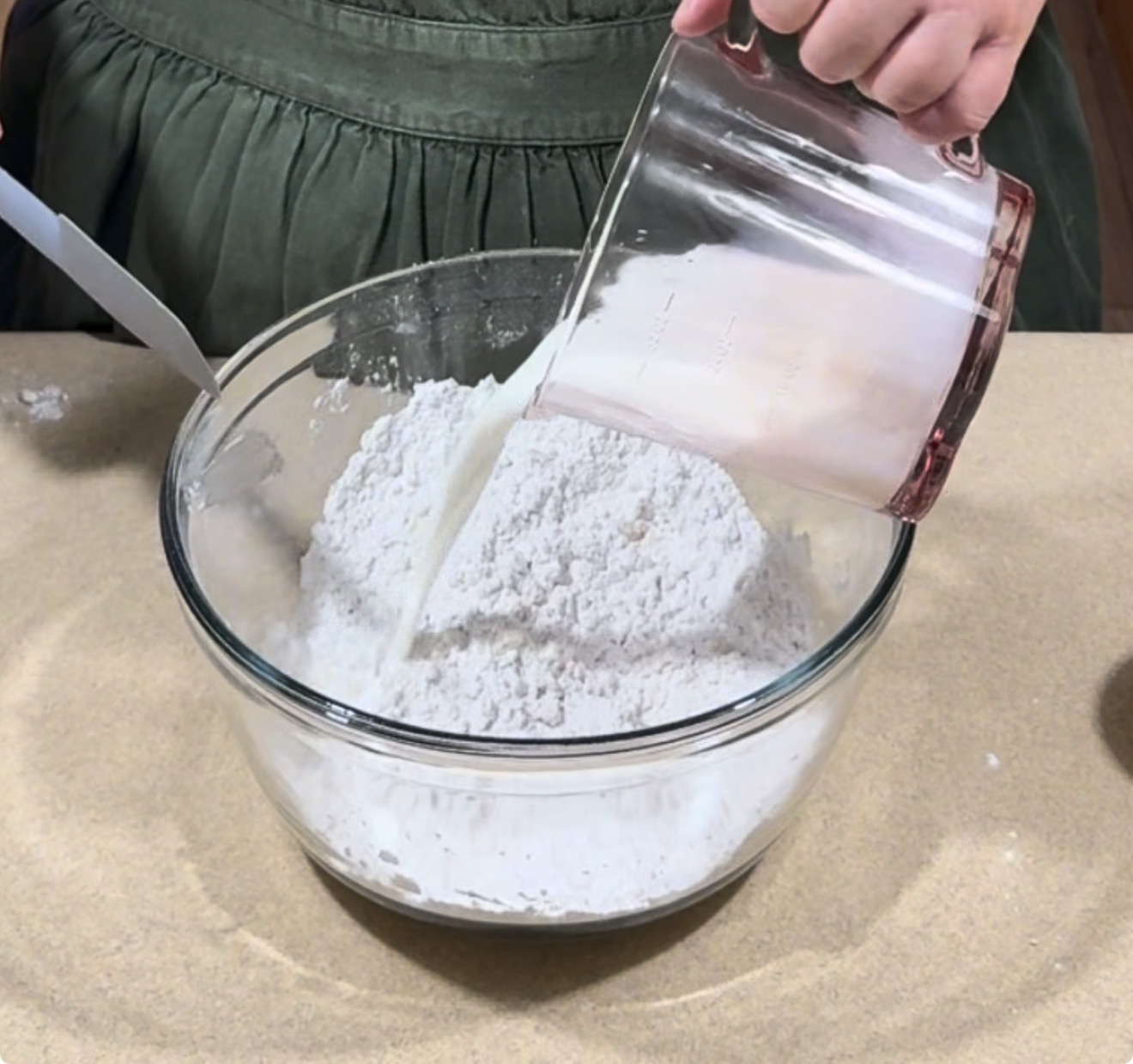 A person pours liquid from a measuring cup into a glass bowl filled with flour on a countertop, preparing ingredients for baking.