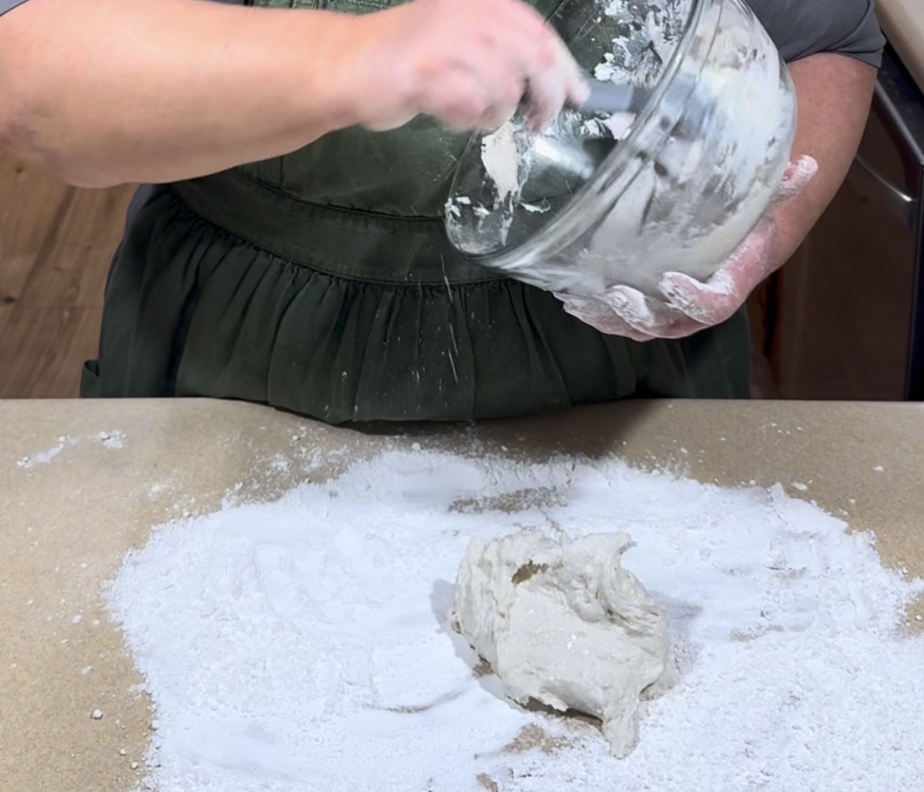 A person in a dark apron scrapes sticky dough from a glass bowl onto a floured surface, preparing for kneading or shaping the dough.