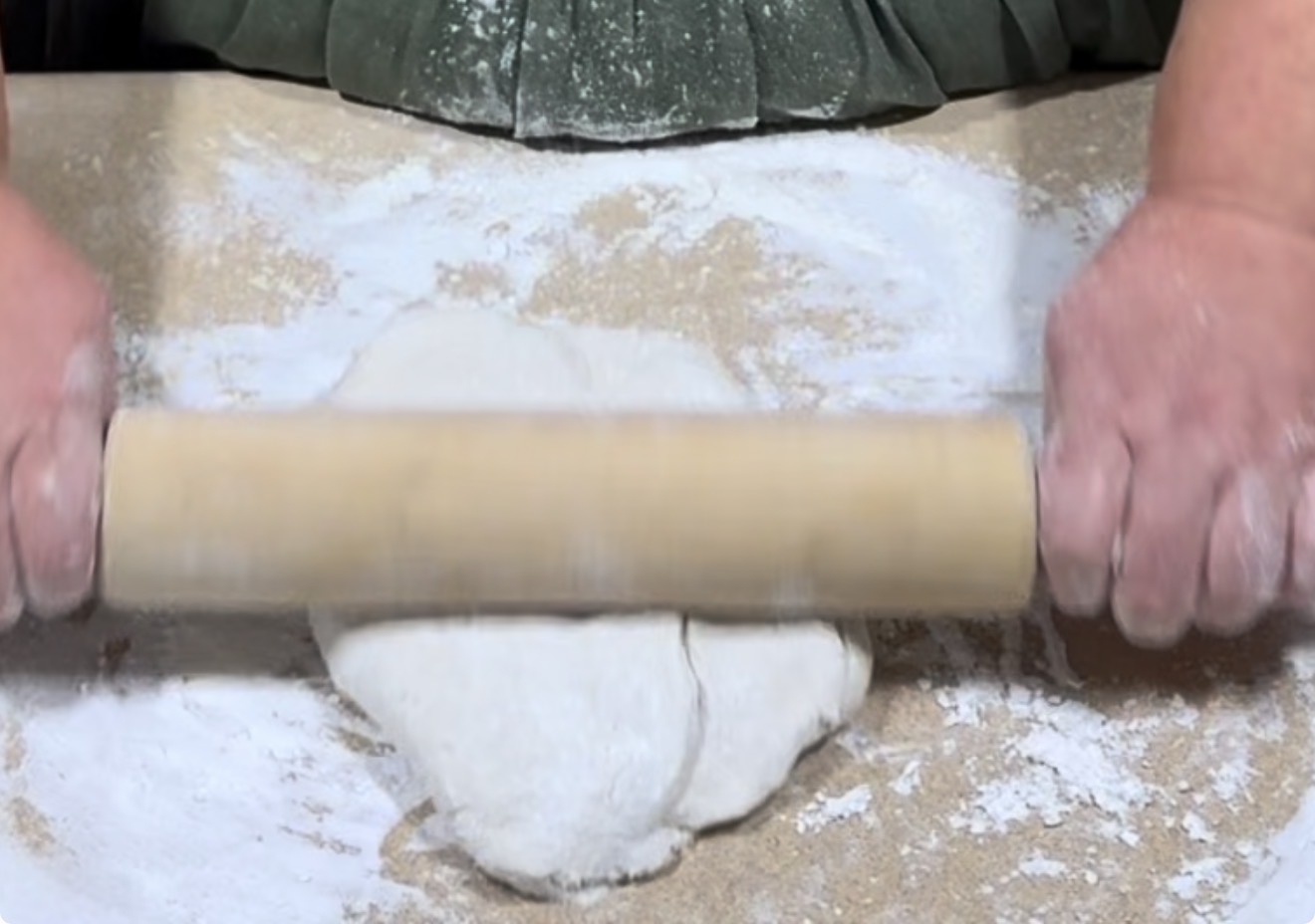 A person uses a wooden rolling pin to flatten dough on a floured surface, preparing it for baking.