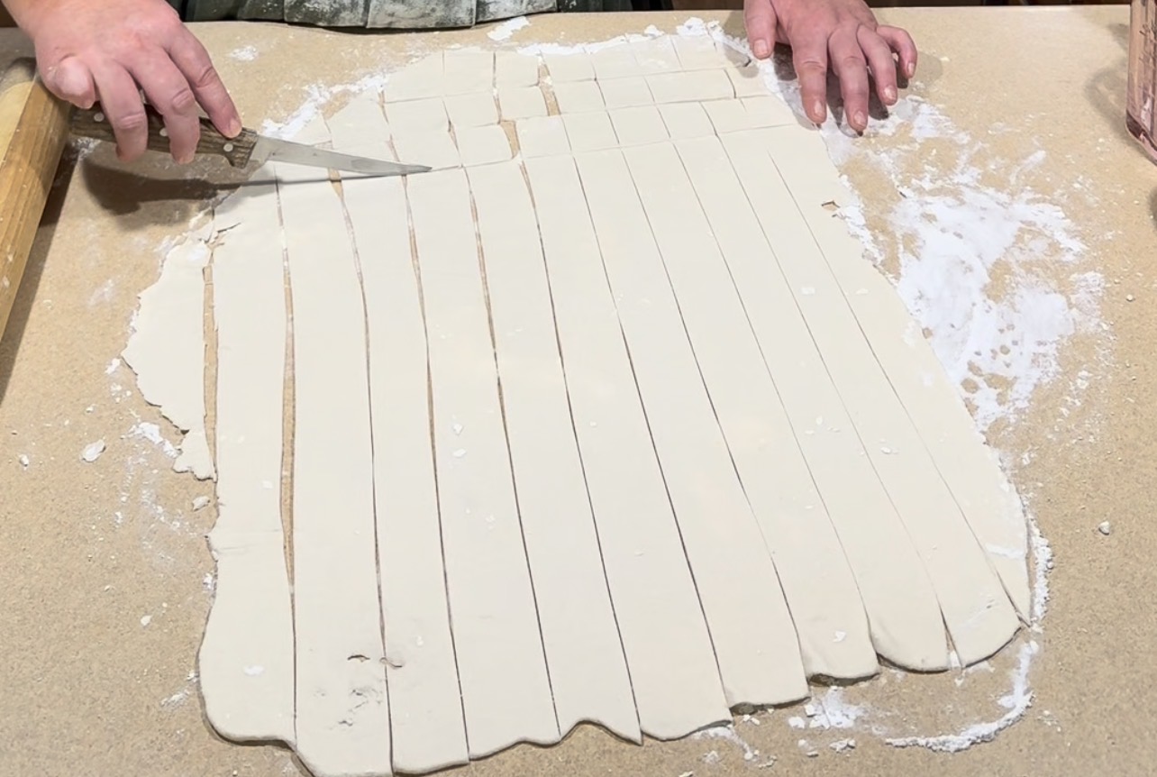 Hands cut long strips of rolled-out dough with a knife on a floured surface. The dough is evenly sliced, and flour is sprinkled on the counter.