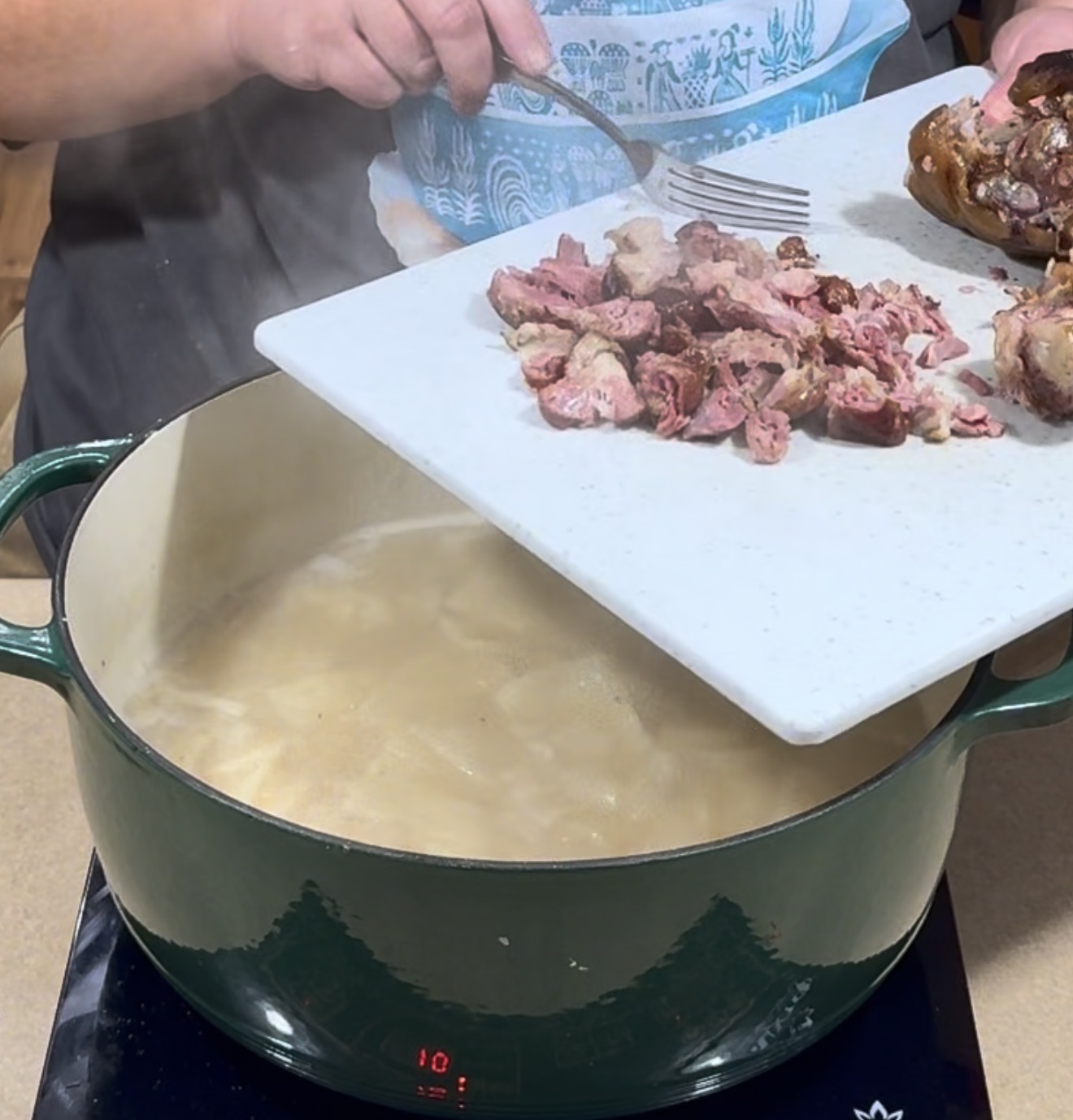 A person adds chopped cooked meat from a cutting board into a steaming green pot of boiling liquid on a stovetop.