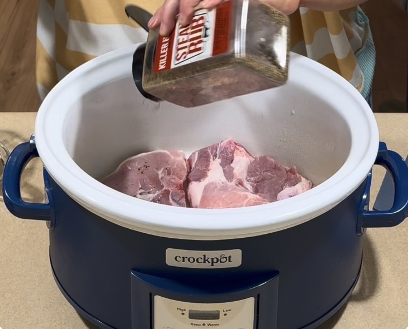 A person sprinkles seasoning onto raw pork in a blue crockpot slow cooker on a kitchen counter.