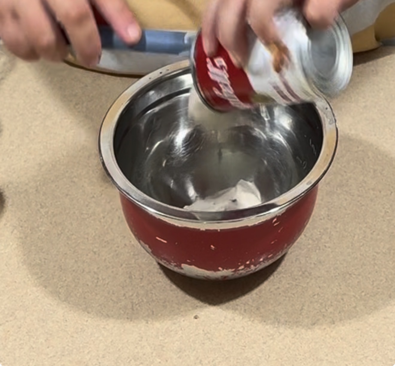 A person pours condensed soup from a can into a red metal mixing bowl on a beige countertop.