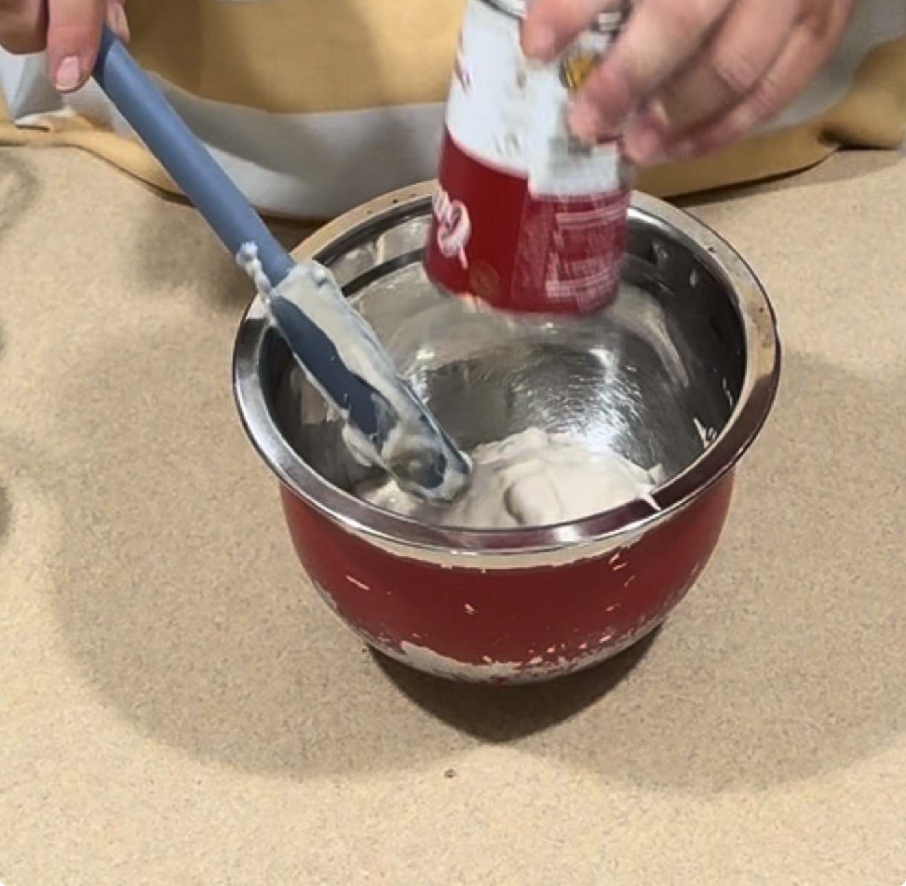 A person scrapes condensed soup from a can into a metal mixing bowl using a blue spatula on a beige counter.