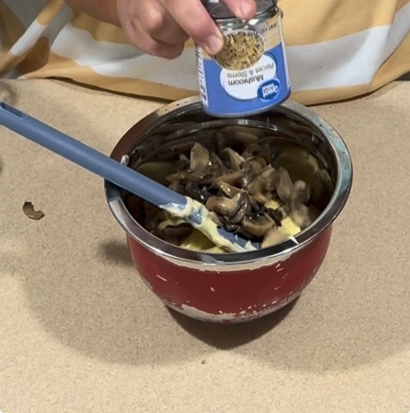 A person pours canned mushrooms into a red mixing bowl filled with sautéed mushrooms and a blue spatula, on a beige countertop.